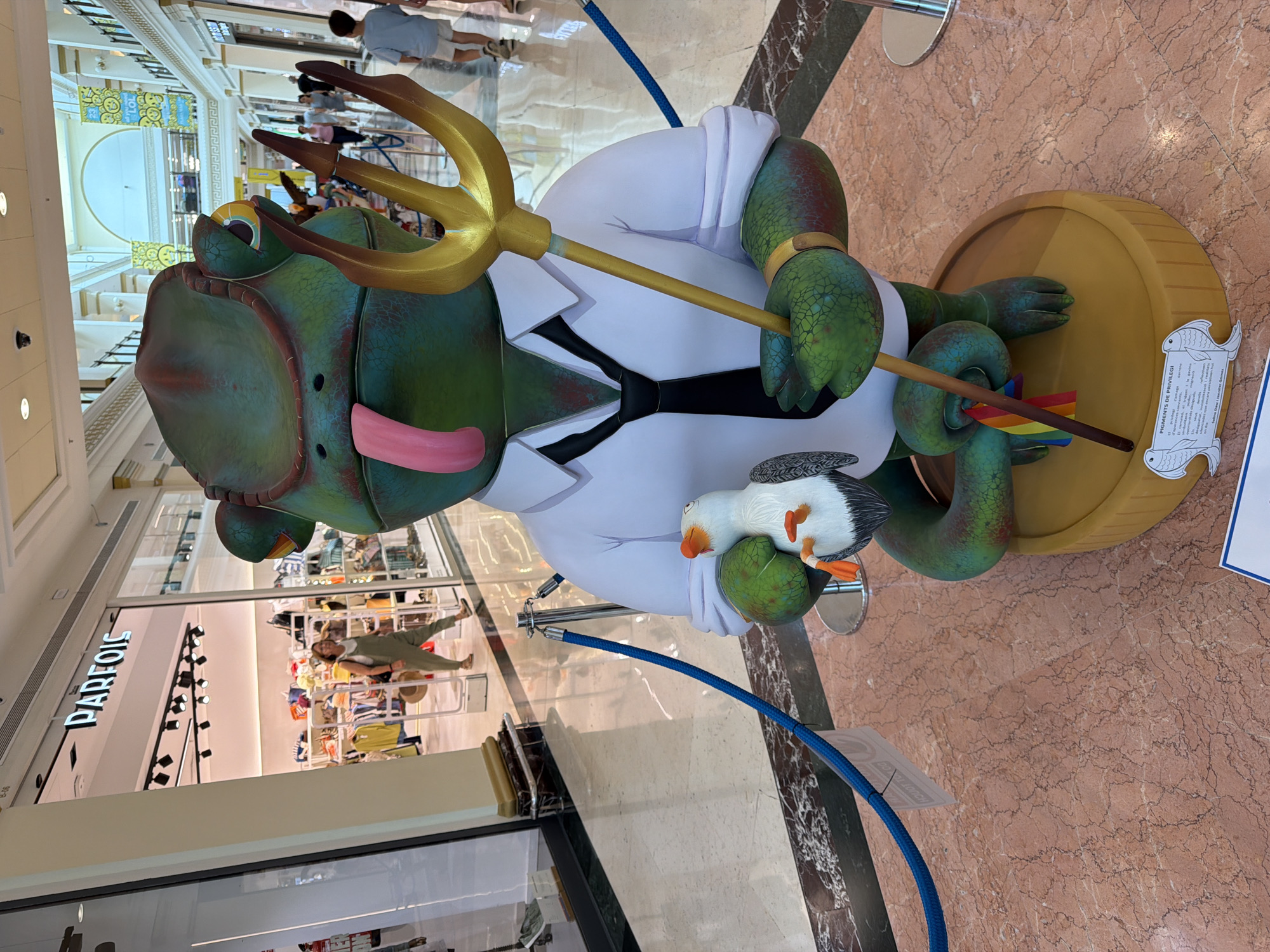This is a medium-shot, eye-level, outdoor photograph taken in a shopping mall in Unknown, Unknown. The focal point is a large, cartoonish, green reptilian statue wearing a white shirt and black tie. The statue has its tongue sticking out, is holding a golden trident in its right hand, and a small seagull figurine in its left hand. A rainbow flag is attached to the trident. The statue is standing on a yellow circular base with a plaque displaying text and two fish drawings. In the background, a woman in green overalls is walking into a store named "PARFOIS". Other shoppers are visible in the distance. The floor is a polished pink marble. Blue ropes on stanchions cord off the area around the statue.