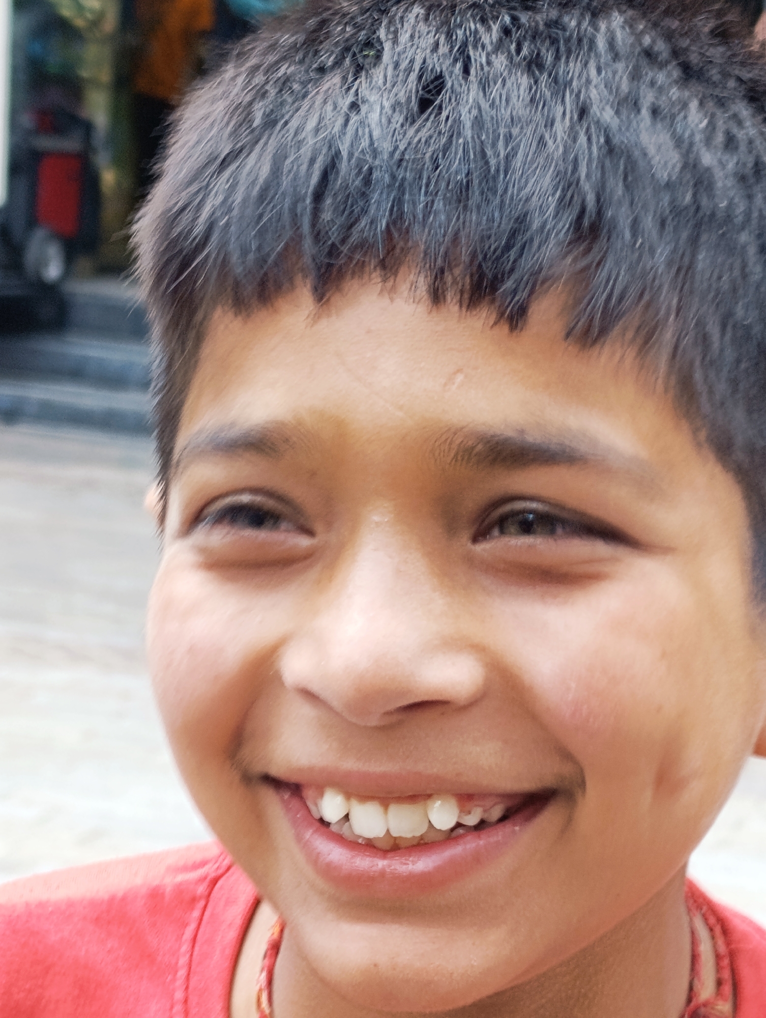 A close-up portrait captures a young boy with dark, tousled hair and fair skin, smiling broadly. His eyes are dark and his cheeks have distinct dimples. He is wearing a red shirt and a beaded necklace. The background is blurred, showing hints of an outdoor environment with what appears to be buildings and possibly steps. The lighting suggests it is daytime. The scene is likely situated in a public area of Manali, India, given the visual cues and context.