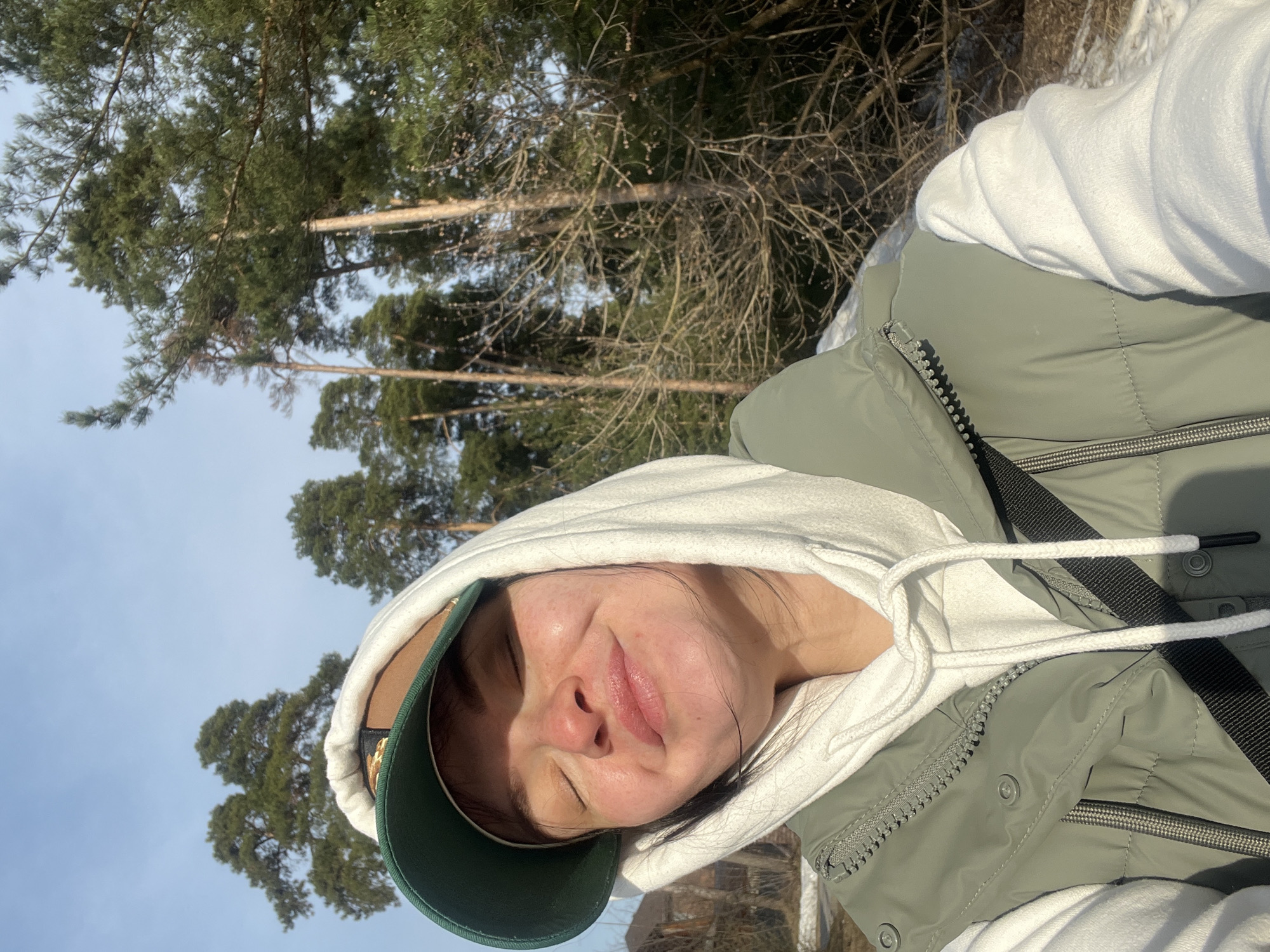 A person is wearing a white hooded sweatshirt, green vest and a green baseball cap. They are outside in a wooded area with tall trees and a blue sky. It is sunny. This person is in Russia, but the picture was not taken in Kazan.