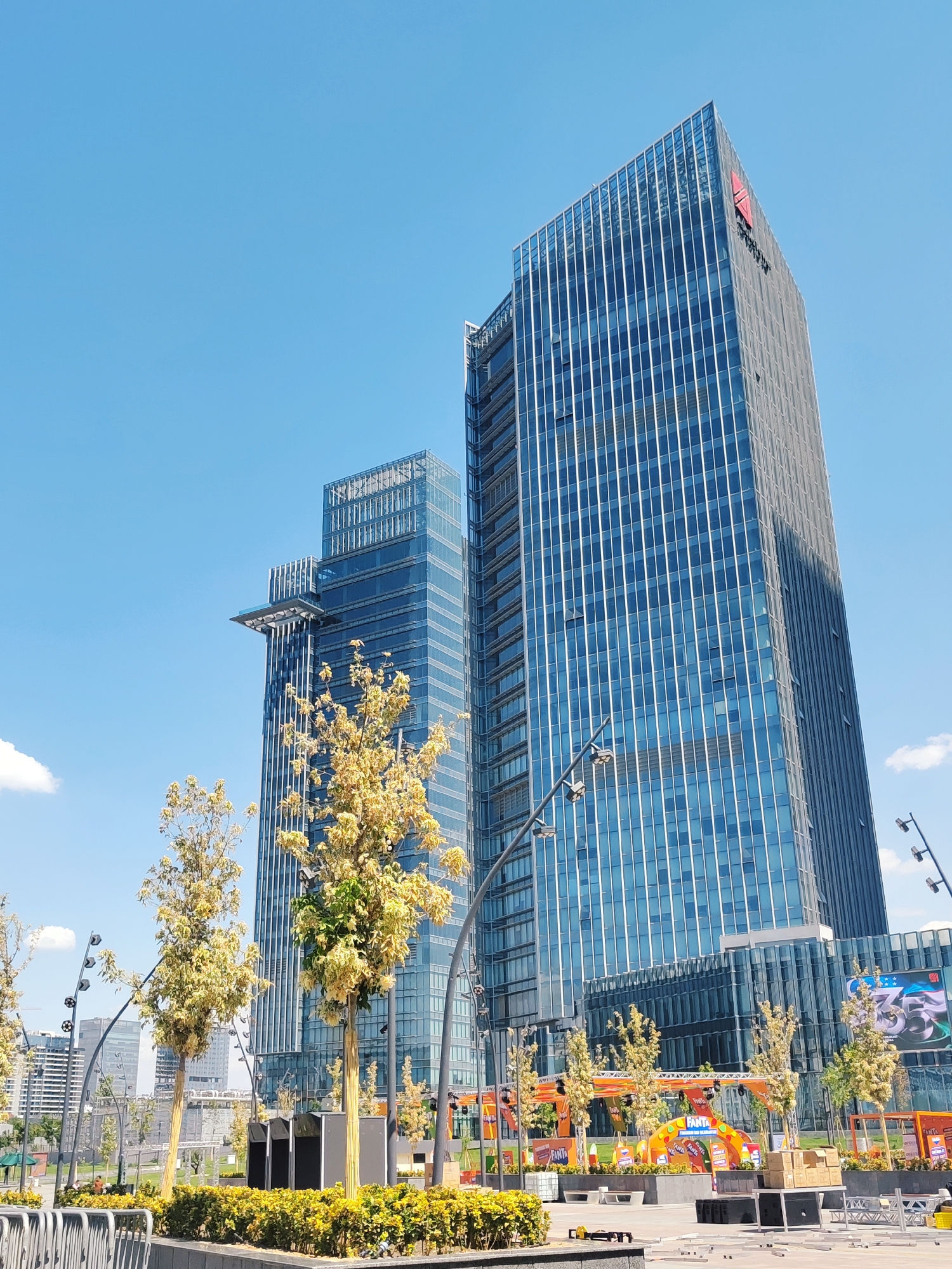 This image depicts the modern business district of Tashkent, Uzbekistan, specifically focusing on the impressive architecture of the Tashkent City complex. In the foreground, several deciduous trees with sparse, pale yellow foliage are visible, with lush yellow-green bushes lining a walkway. Modern streetlights with multiple spherical fixtures are strategically placed throughout the scene. Behind the trees, two towering glass-facade skyscrapers dominate the skyline. The taller skyscraper on the right features a prominent red logo with a stylized "A" at its apex, and vertical white lines accentuating its glass exterior. The second skyscraper to the left has a distinctive cantilevered platform extending from its upper section. In the middle ground, an outdoor event setup is visible, characterized by colorful arched structures and banners promoting "FANTA" beverages. A large LED screen displays graphics, including the number "355". Beyond the skyscrapers, other buildings and urban infrastructure are discernible under a clear blue sky with scattered white clouds. The scene conveys a sense of urban development and contemporary city life.