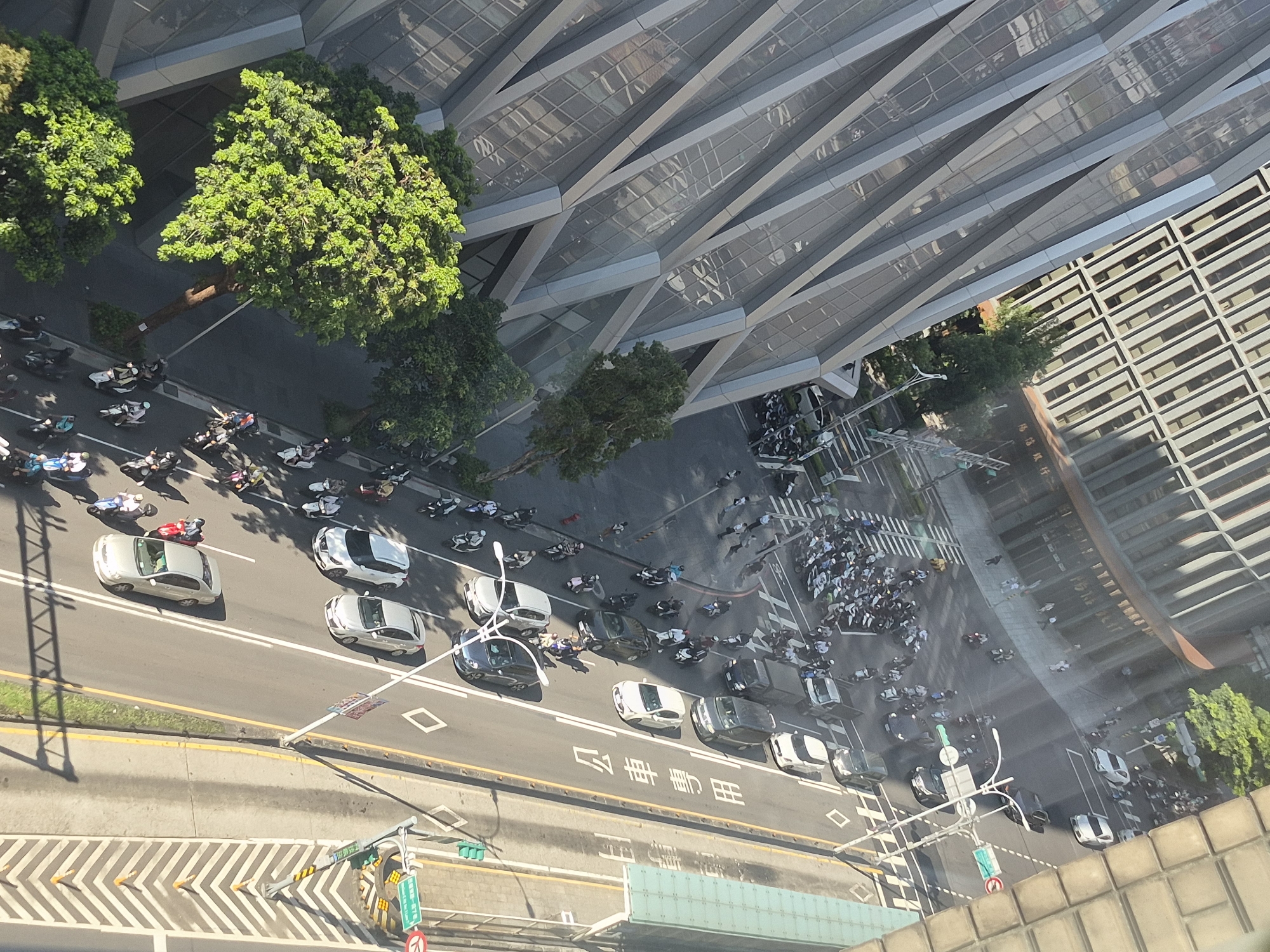 The aerial photograph, taken from a high vantage point, captures a sunlit street scene in Taipei, Taiwan, likely during daytime, given the clear shadows. The dominant structures are a modern, angular glass-fronted building on the left, reflecting the sky and surrounding environment, and a multi-story building with a patterned facade in the background.
A busy street runs diagonally through the frame, characterized by multiple lanes of traffic. The street is populated by a significant number of scooters and several cars. Scooters are the predominant mode of transport visible, with many parked closely together in designated areas and others in motion. Cars include sedans and a few vans. Pedestrians are present on the sidewalks and crossing the street, some walking and others on scooters.
The road is marked with white lines, including a double yellow line separating lanes and diamond-shaped markings. Traffic signals are visible, indicating active traffic control. Green trees are strategically placed along the street and near the buildings, adding a natural element to the urban landscape. The overall impression is one of a bustling city environment with a high density of two-wheeled vehicles. Chinese characters are visible on a sign on the right side of the road, confirming the location as Taiwan.