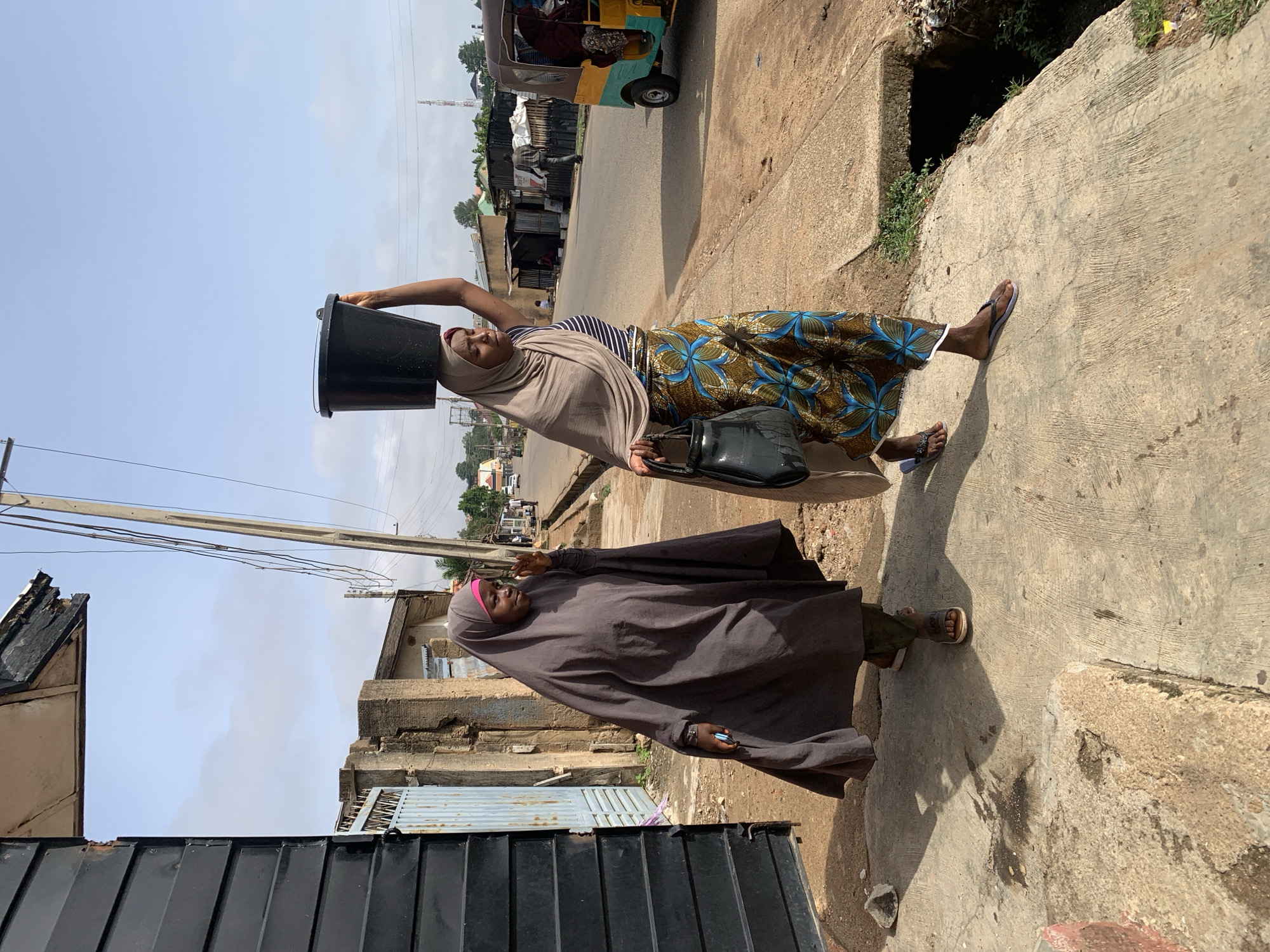 Two women walk along a concrete pathway in an urban or semi-urban street scene, likely in Jos, Nigeria, during the daytime under a clear, bright sky. The image is taken from a slightly low angle, looking upwards towards the sky.
The woman in the foreground (left side of the image if upright) wears a dark brown abaya or loose garment, covering her head with a similar-colored hijab and possibly a pink underscarf. She has light-colored sandals on her feet.
Behind her, another woman walks, balancing a black bucket on her head with her right hand. She wears a beige head covering, a striped top (partially visible), and a long, vibrant skirt or wrapper with blue and brown floral patterns. She carries a black handbag in her left hand and wears purple flip-flops.
The women are walking on a concrete path beside an open drainage channel or ditch. In the background, a paved road is visible, with a yellow and green tricycle (keke napep) further down the street. Several buildings, some appearing residential and others with corrugated metal roofs and gates, line the street. Power lines and utility poles are also present against the sky. No visible text is discernible in the image. The scene suggests a typical daily activity in the specified region.