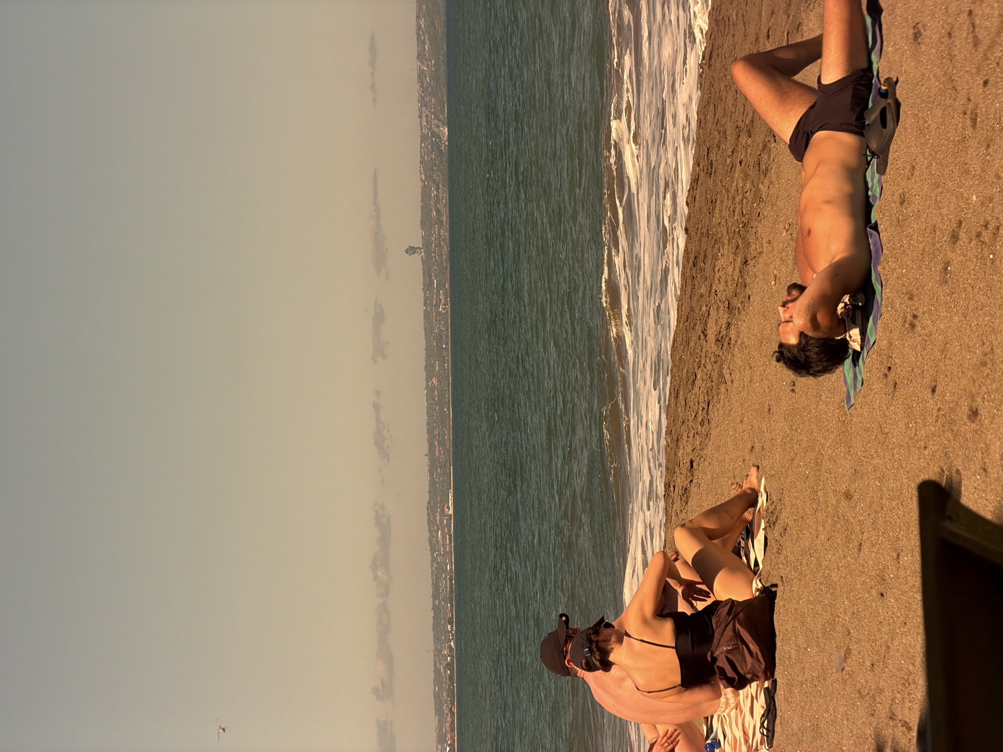 The image depicts a beach scene with three people relaxing on the sand. The location is likely in or near a coastal area, given the presence of the ocean and sandy shoreline.
On the right side of the image, a person is lying on their back, close to the water's edge. They appear to be sunbathing, and a towel is visible beneath them. Another person is lying next to a woman with dark hair. The woman is wearing a black bathing suit top and a baseball cap.
The environment consists of a beach with a sandy shoreline meeting the ocean. The water has visible waves. The sky appears to be clear. The location is near North Kuta, Indonesia.