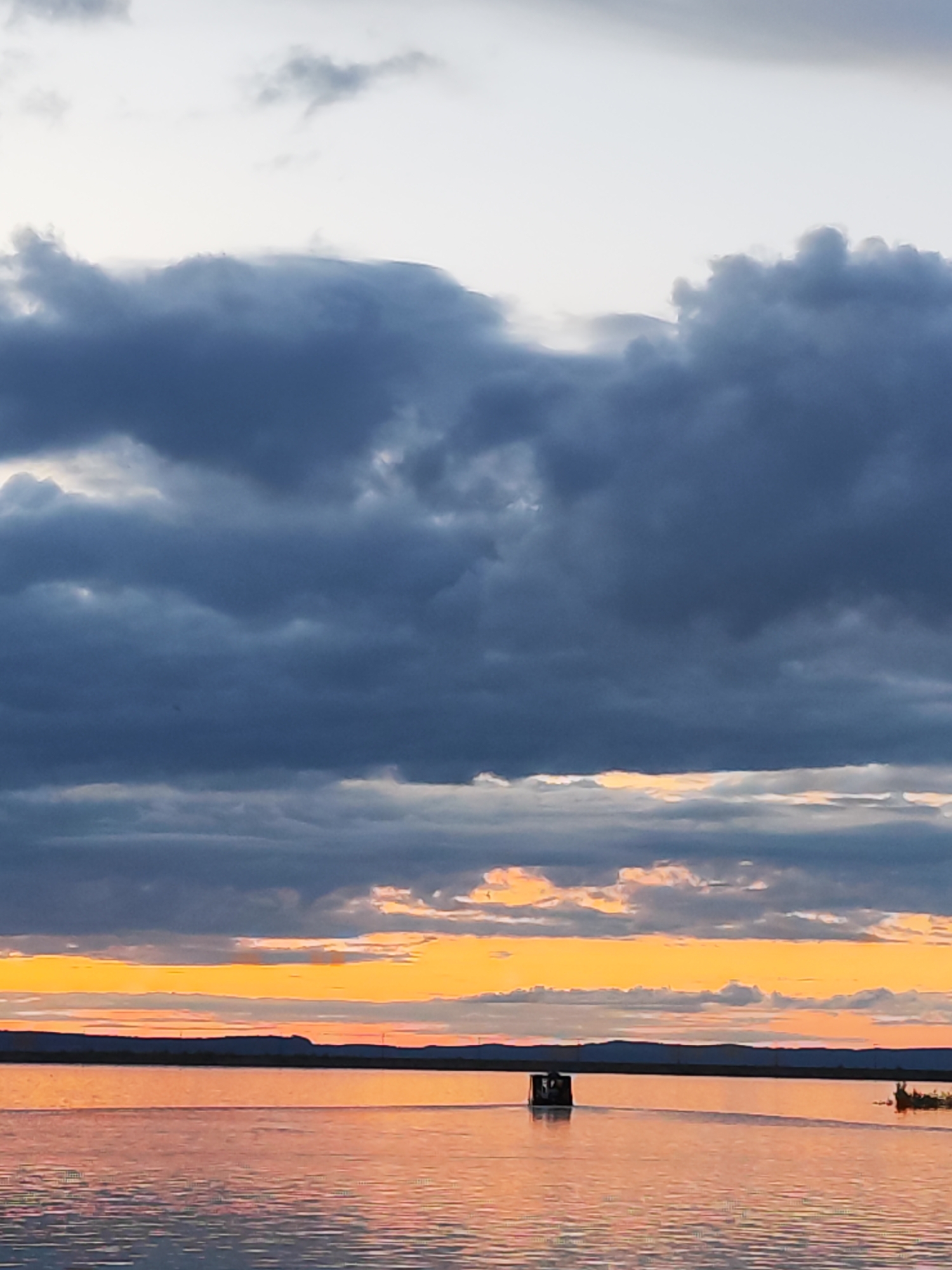 The image shows a sunset over the Kama River in Chaikovsky, Russia. The sky is filled with layers of dark, heavy clouds above a band of orange and yellow light near the horizon. The river reflects the colors of the sunset, creating a warm, shimmering effect on the water's surface. There is a small, box-like structure on the water, possibly a floating platform or small building, and a smaller object to the right, perhaps a small boat or patch of reeds. In the distance, a line of land is visible.