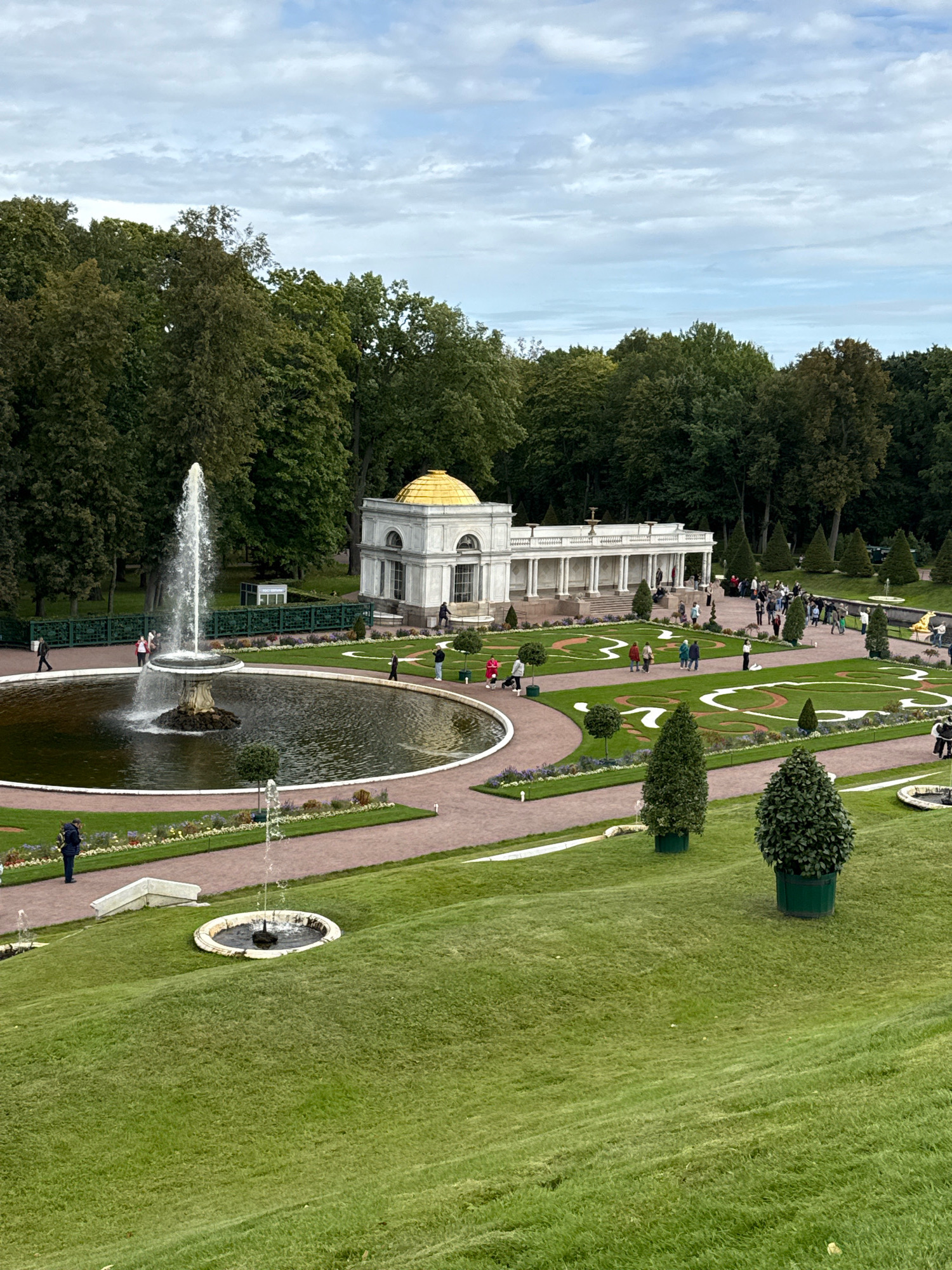 This image depicts a view of the Peterhof Palace complex in Peterhof, Russia, specifically focusing on the Grand Cascade area. In the foreground, a large circular fountain with a central sculpture erupts with a tall spray of water, surrounded by manicured lawns and pathways. Several smaller fountains are visible in the midground. A white classical-style building with a golden dome and a colonnaded facade is situated behind the main fountain. Numerous people are scattered throughout the scene, walking along the paths and observing the fountains and architecture. The background consists of a dense line of mature trees, with a cloudy sky overhead. The overall environment is an elaborate formal garden with a prominent water feature and architectural elements.