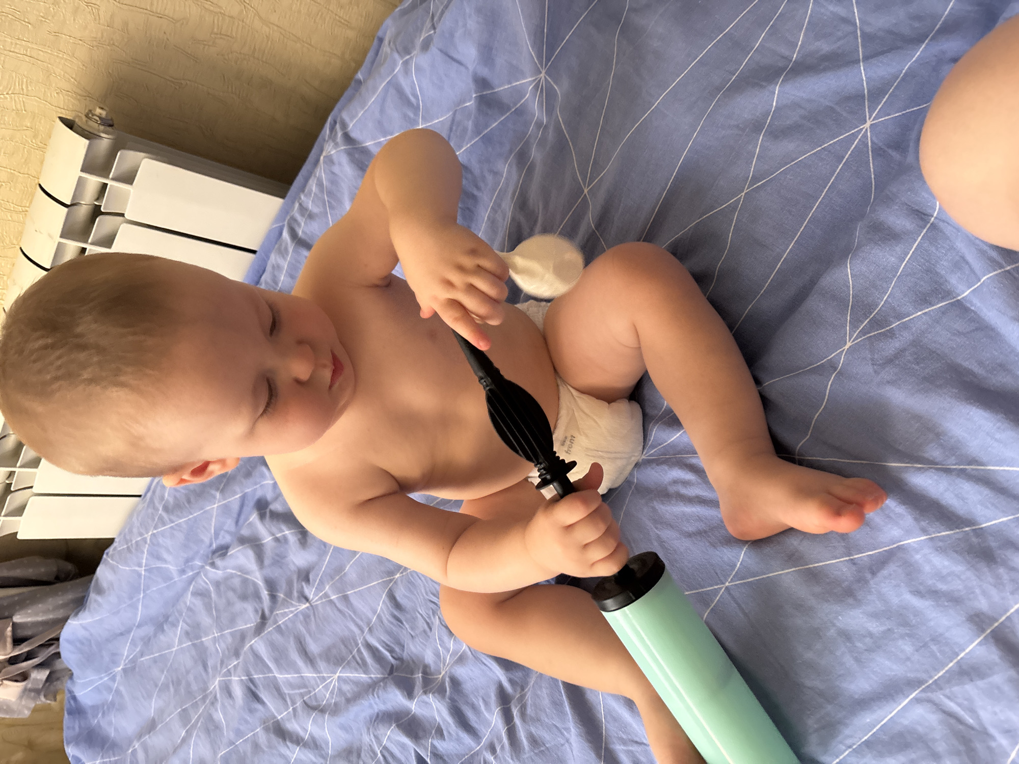 A baby is sitting on a blue-patterned sheet, likely a bed or mat. The baby is wearing a diaper and appears to be around one year old, with light brown hair and closed eyes. The baby is holding a light green pump with a black nozzle, appearing to be a balloon pump, with their hands. Another object, a white scoop or bowl-like item, is near the baby's chest.
In the background, to the left, is a white radiator with black accents and a textured beige wall. The lighting suggests daytime. The overall impression is one of a quiet, indoor moment. No text is visible in the image.
