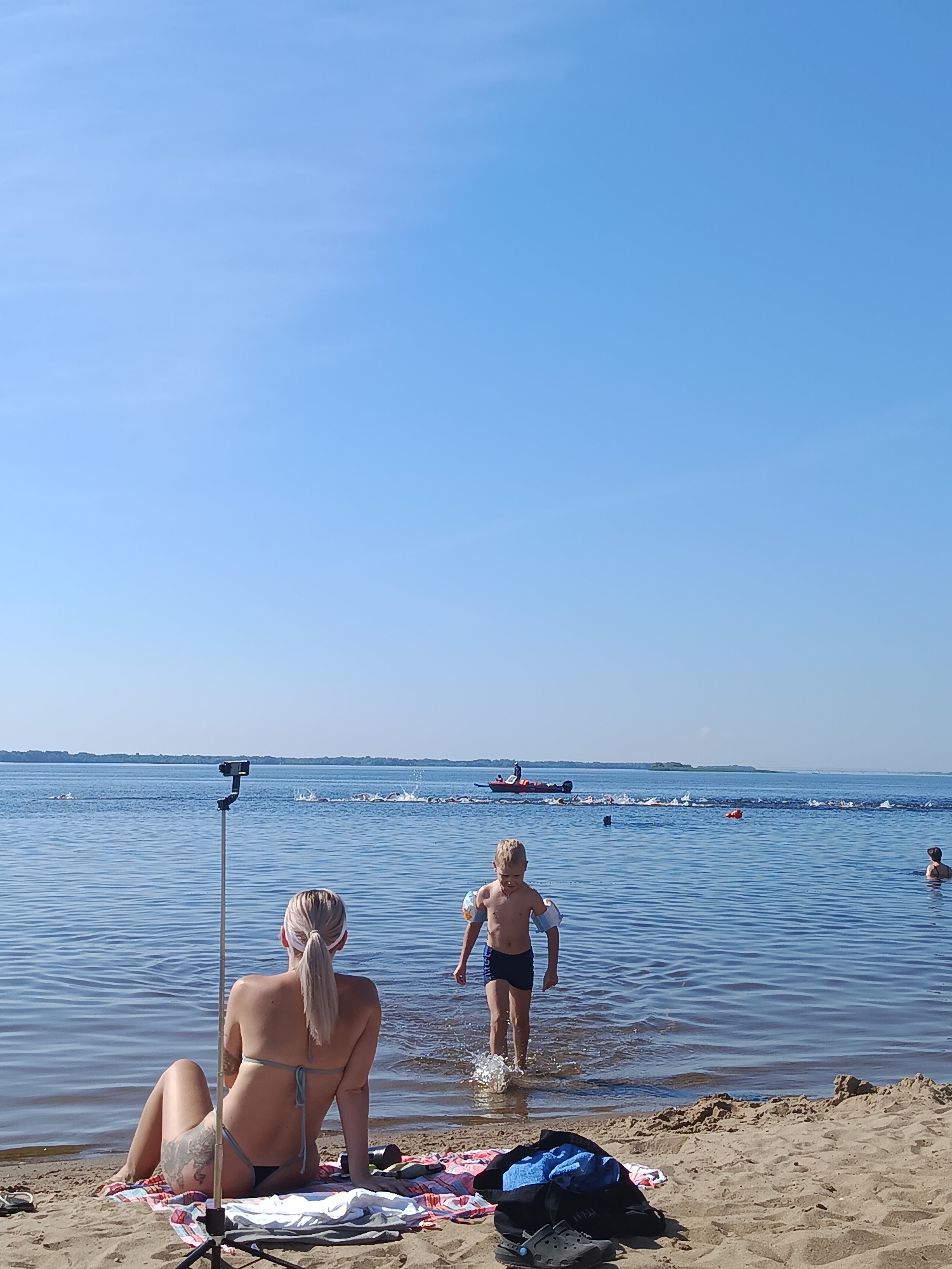 This image depicts a sunny day at a body of water, likely a river or lake, with a beach in the foreground. A woman with blonde hair tied in a ponytail, wearing a bikini, is sitting on a patterned blanket on the sand, facing away from the camera. A selfie stick is positioned in front of her. Further into the water, a young boy wearing swim trunks and arm floaties is walking into the water, splashing. In the middle ground, a group of people are swimming in a line, marked by buoys, with a red boat with people on it nearby. In the distance, a shoreline with trees is visible under a clear blue sky. The scene suggests a recreational outing, possibly a swimming competition or training event. The image does not contain any specific landmarks that would allow for definitive identification of the location within Saratov, Russia, but the open water, beach, and distant treeline are consistent with areas along the Volga River.