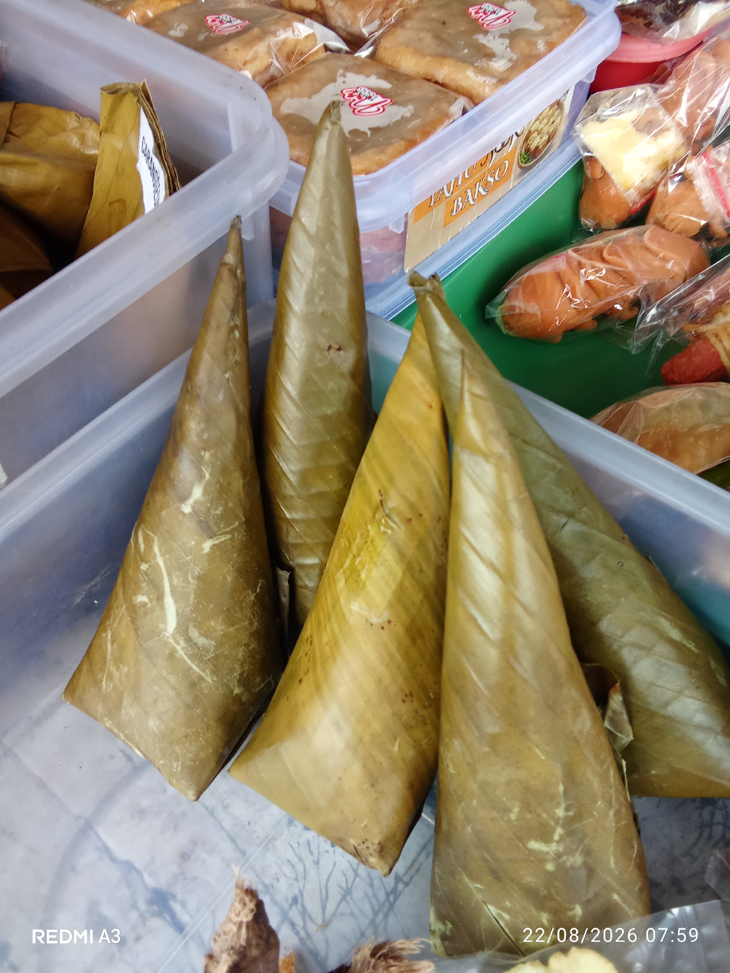 The image shows a close-up of several cone-shaped food items wrapped in brown leaves, likely a local Indonesian delicacy, arranged in a transparent plastic container. In the background, other packaged food items are visible in similar containers and plastic wrapping. The scene appears to be a street food stall or market display. The timestamp "22/08/2026 07:59" is visible in the lower right corner, suggesting the image was taken on August 22, 2026, at 7:59 AM. The text "REDMI A3" is also present in the lower left, indicating the phone model used to capture the image. The setting is likely outdoors or in a covered market, given the arrangement of goods for sale. There are no people visible in the frame.