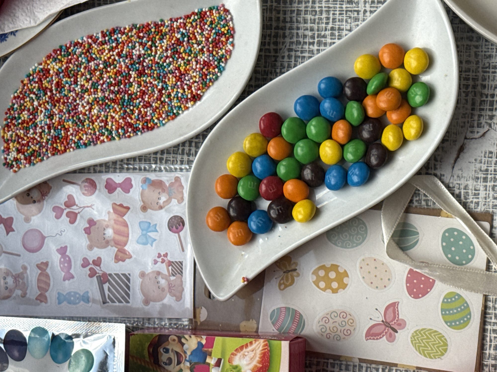 The image shows two white, irregularly shaped plates on a grey textured surface. The plate on the left contains multicolored sprinkles covering most of its area. The plate on the right contains M&M candies in various colors including yellow, orange, green, blue, red, and brown. Below the plates are various items including sticker sheets featuring teddy bears, easter eggs, and butterflies. There is also a partial view of a small cardboard box. The image was taken indoors, likely in a residential setting. There is no evidence to confirm this photo was taken in Yerevan, Armenia.