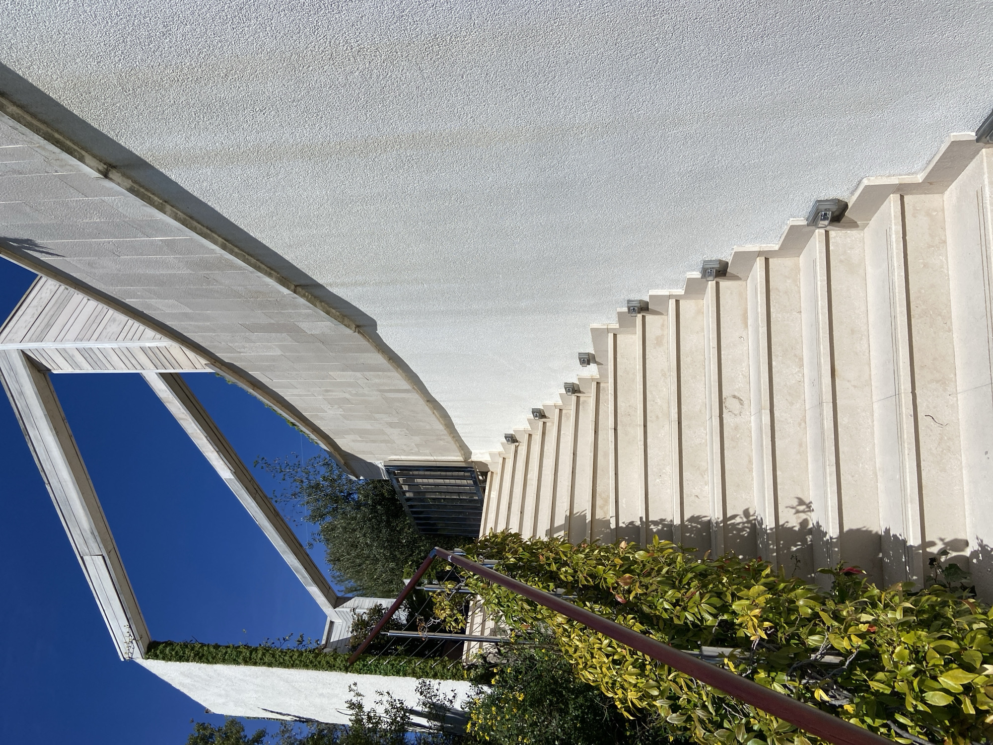 The image shows an upward-angled view of a building exterior, likely located in a sunny climate. The sky is bright blue, visible through the angled structural supports of a covered walkway or awning. The building itself is light-colored with a textured surface. A series of evenly spaced, rectangular panels or columns run vertically, each capped with a small, dark lighting fixture. Green vegetation, including climbing plants and bushes, is growing along the base of the building and near a red metal railing. The overall impression suggests a modern or contemporary architectural style set within a garden-like or landscaped area. The location is stated to be in Budva, Montenegro, but without further information, it is impossible to locate the building to a more specific location.