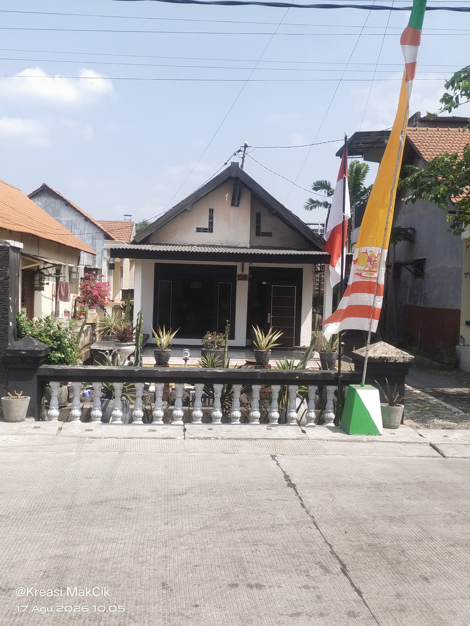 The image depicts a residential street scene in Sambi Bulu, Indonesia. In the foreground, a concrete road with parallel lines runs horizontally across the frame. A low, white, ornate fence with a black base runs along the edge of a property. Beyond the fence, a collection of potted plants, including cacti and other succulents, are arranged. A house with a dark-colored entrance and a white facade is visible behind the plants and fence. The house has a dark-colored roofline with visible framing. Two flags are prominent: one is red and white, possibly the Indonesian flag, and the other is a long yellow and orange flag with decorative elements. The sky is blue with scattered clouds. The overall environment suggests a typical residential area in Indonesia. The timestamp at the bottom left corner indicates "17 Agu 2026 10.05" and a watermark "@Kreasi MakCik".