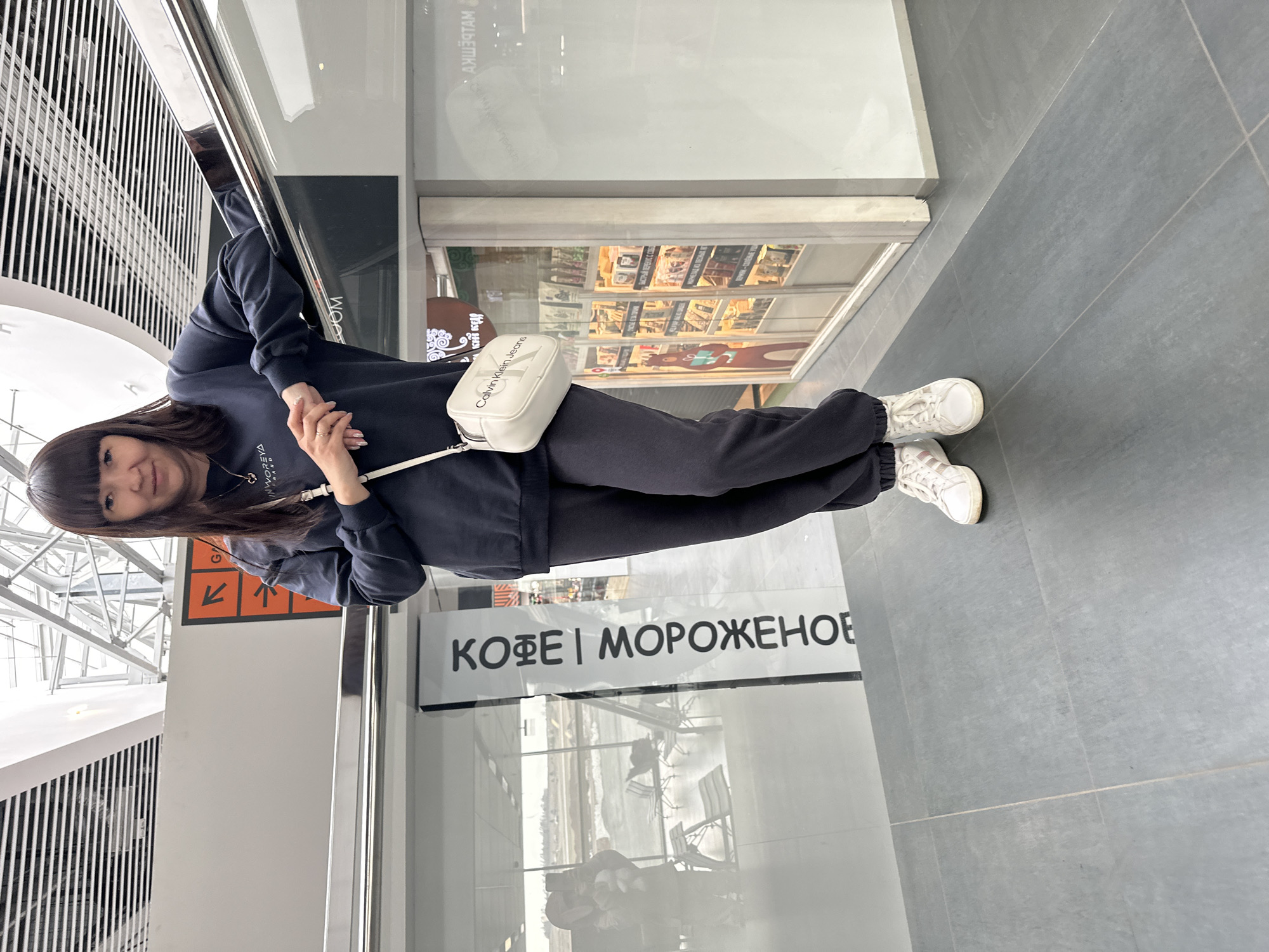 A woman with shoulder-length dark hair and bangs is standing in an indoor location, likely a shopping mall or transportation hub. She is wearing a dark blue sweatshirt, dark sweatpants, and white sneakers. She is also carrying a small, white Calvin Klein crossbody bag. She is standing in front of a glass partition with the words "КОФЕ | МОРОЖЕНОЕ" (Coffee | Ice Cream) printed on it in Russian. There are orange signs visible on the left and the environment suggests the location is the city of Bolshoye Savino, Russia. The floor is made of gray tiles. The woman appears to be posing for the camera.