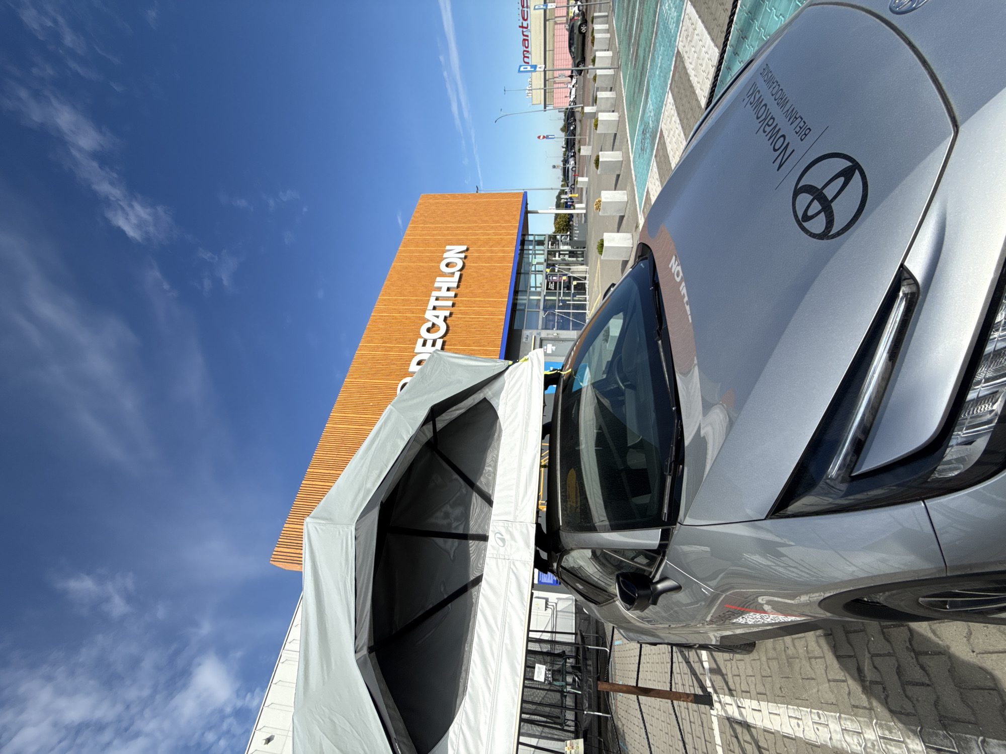 An outdoor, medium-shot, eye-level, wide-angle photograph captures a silver Toyota car parked in a brightly lit outdoor parking lot. The car is equipped with an open, light gray rooftop tent, which covers the majority of the vehicle's roof and extends slightly over the windshield. The car's hood displays the Toyota logo and the text "Nowakowski Bielany Wrocławskie". The parking lot features a distinctive checkered pattern of green and white painted areas. In the background, a large, modern building with an orange, slatted facade stands prominently, with the word "DECATHLON" emblazoned across it in large white letters. To the right of the Decathlon building, another store with a sign that reads "pmartes" is visible, along with additional parking spaces and other vehicles. The sky above is a clear, vibrant blue with scattered white clouds. The scene appears to be in the city of Wrocław, Poland, likely at a retail or commercial area. No people are visible in the frame.