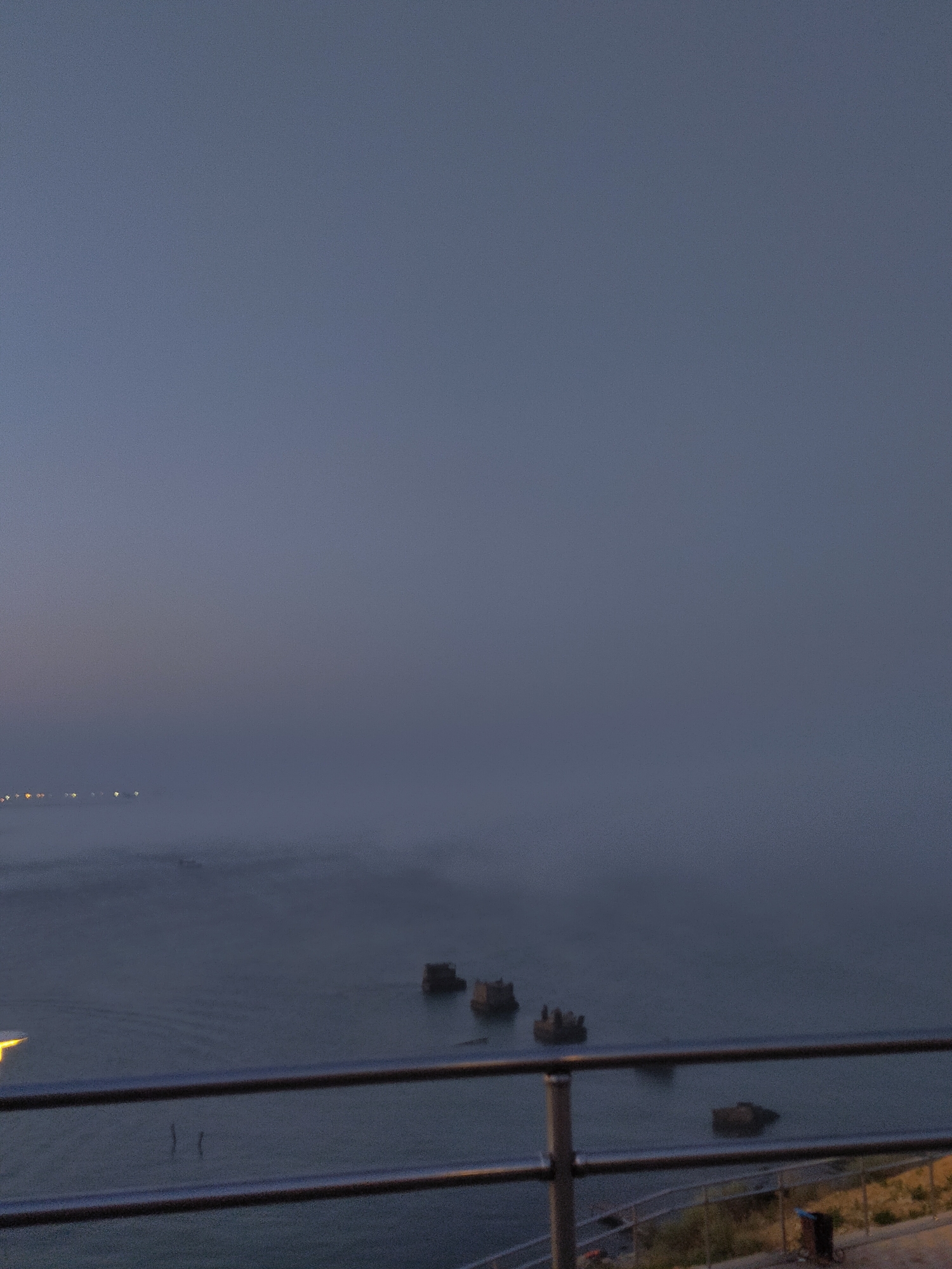 The image depicts a foggy evening scene overlooking the Caspian Sea in Aktau, Kazakhstan. In the foreground, a metal railing runs horizontally across the frame, suggesting a vantage point from a walkway or balcony. Beyond the railing, the sea is visible, shrouded in mist. Several dark, rectangular structures, possibly remnants of old ships or barges, are partially submerged in the water. In the distance, faint lights from the city of Aktau are visible through the fog, creating a hazy glow. The sky is a gradient of deep blue to a lighter, muted grey, characteristic of twilight or dawn. The overall atmosphere is serene and somewhat melancholic due to the fog and dim lighting. No people are visible in the image.