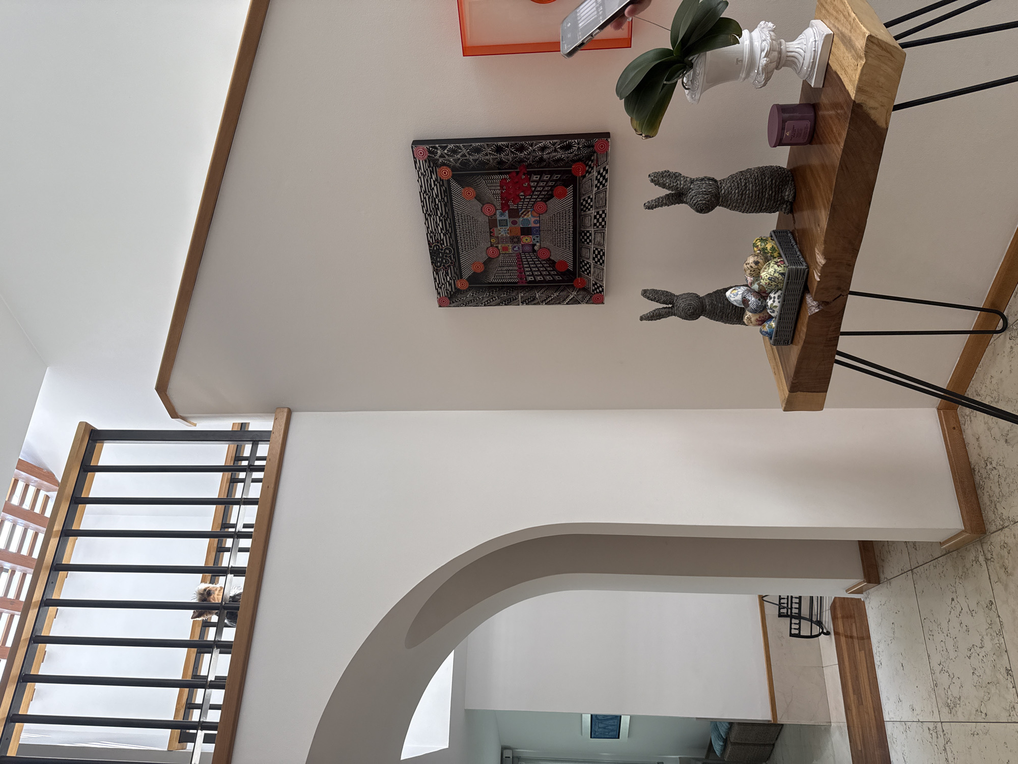The image shows an interior space with a staircase, an arched doorway, and a display shelf. The staircase has wooden railings and black metal bars. An arched doorway leads to another room. On the wall, there's a square artwork featuring geometric patterns and a wooden shelf displaying decorative objects including a white urn with an orchid, two wicker bunnies, and a basket of Easter eggs. The floor is tiled. The location of San Bernardino Tlaxcalancingo, Mexico is not evident from the image.
