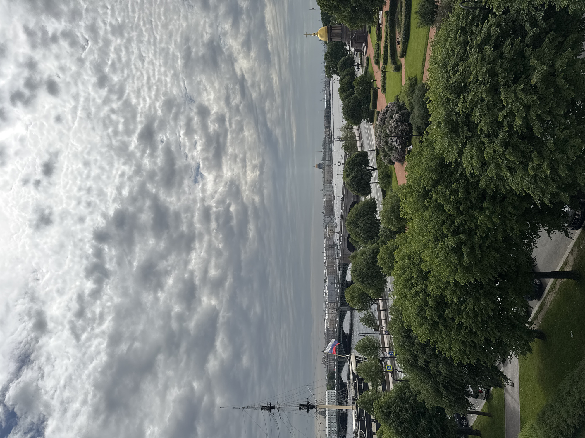 The image captures a panoramic view of Saint Petersburg, Russia, on a cloudy day. The foreground is dominated by lush green trees and manicured park areas with pathways and low hedges. A prominent golden-domed structure, likely a chapel or small church, stands to the right.
In the middle ground, a wide river, the Neva, separates the park from the cityscape. Several bridges span the river, with traffic visible on them. On the far bank, a dense collection of historic buildings forms the city skyline, characterized by various architectural styles. A large sailing ship, possibly a museum ship, is moored on the left side of the river, its masts and rigging clearly visible against the sky. A Russian flag flies from a pole on the ship.
The sky is filled with an intricate pattern of grey and white clouds, creating a dramatic and overcast atmosphere. The overall scene depicts a blend of nature, urban architecture, and historical maritime elements characteristic of Saint Petersburg. No individuals are clearly discernible in the image, but implied activity can be seen in the presence of vehicles on the bridges and along the riverbank.