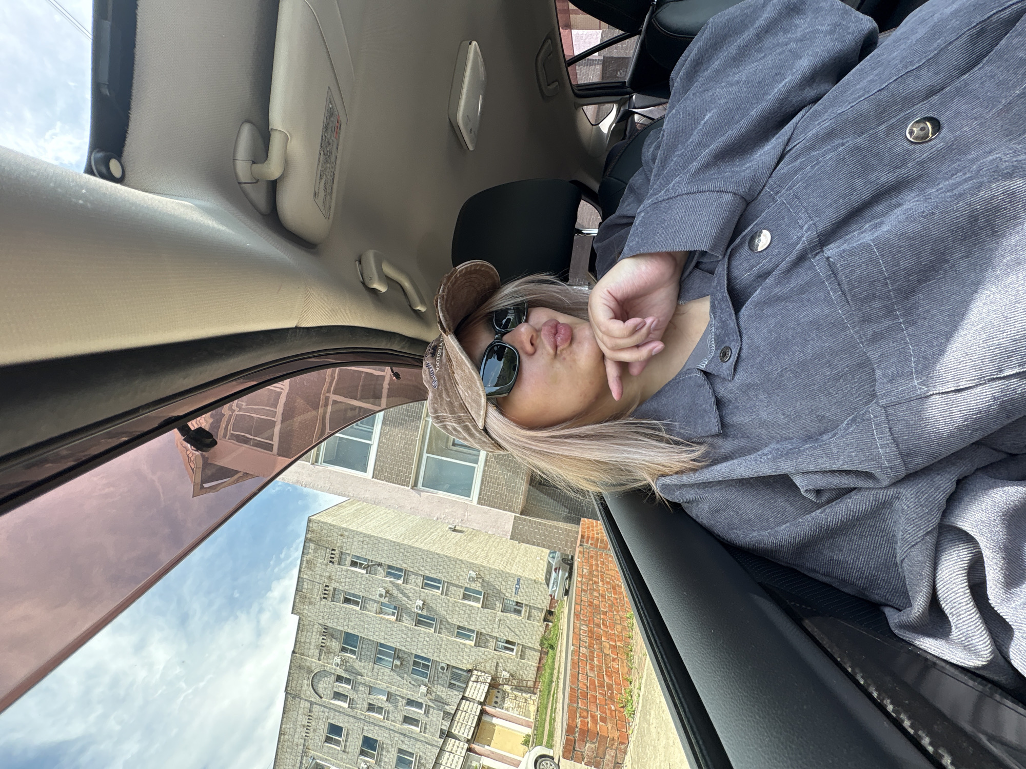 A young woman with blonde hair, wearing a grey corduroy jacket, a brown baseball cap, and sunglasses, sits in the passenger seat of a car. She is puckering her lips and resting her chin on her hand, looking out the window. Outside the car, tall, light-colored apartment buildings with many windows are visible, along with a brick wall and a street with parked cars. The sky is partly cloudy. The scene appears to be in an urban environment, possibly Birobidzhan, Russia, given the architectural style of the buildings.