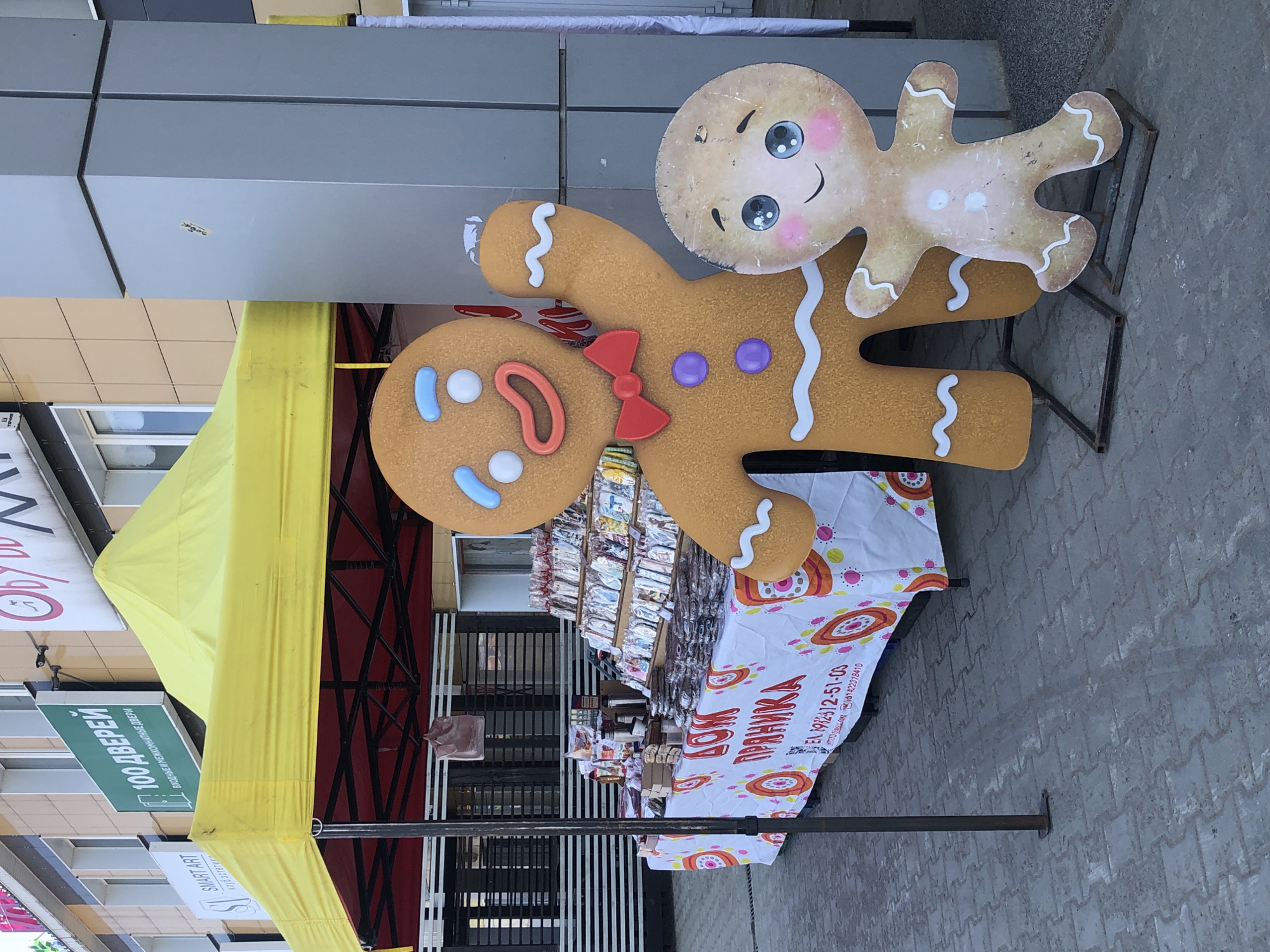 The image captures a street scene in Chaikovsky, Russia, showcasing a small market stall under a yellow canopy. The stall is selling "Дом Пряника" (Gingerbread House) and has gingerbread-themed decorations, including two large gingerbread cutout figures, one holding a smaller one. The stall has a display of goods under a white tablecloth with colorful, circular patterns. There are signs nearby, including one that reads "100 ДВЕРЕЙ" (100 Doors) and another for "SMART ART" ЭСТЕТИК. The stall is situated on a paved walkway.