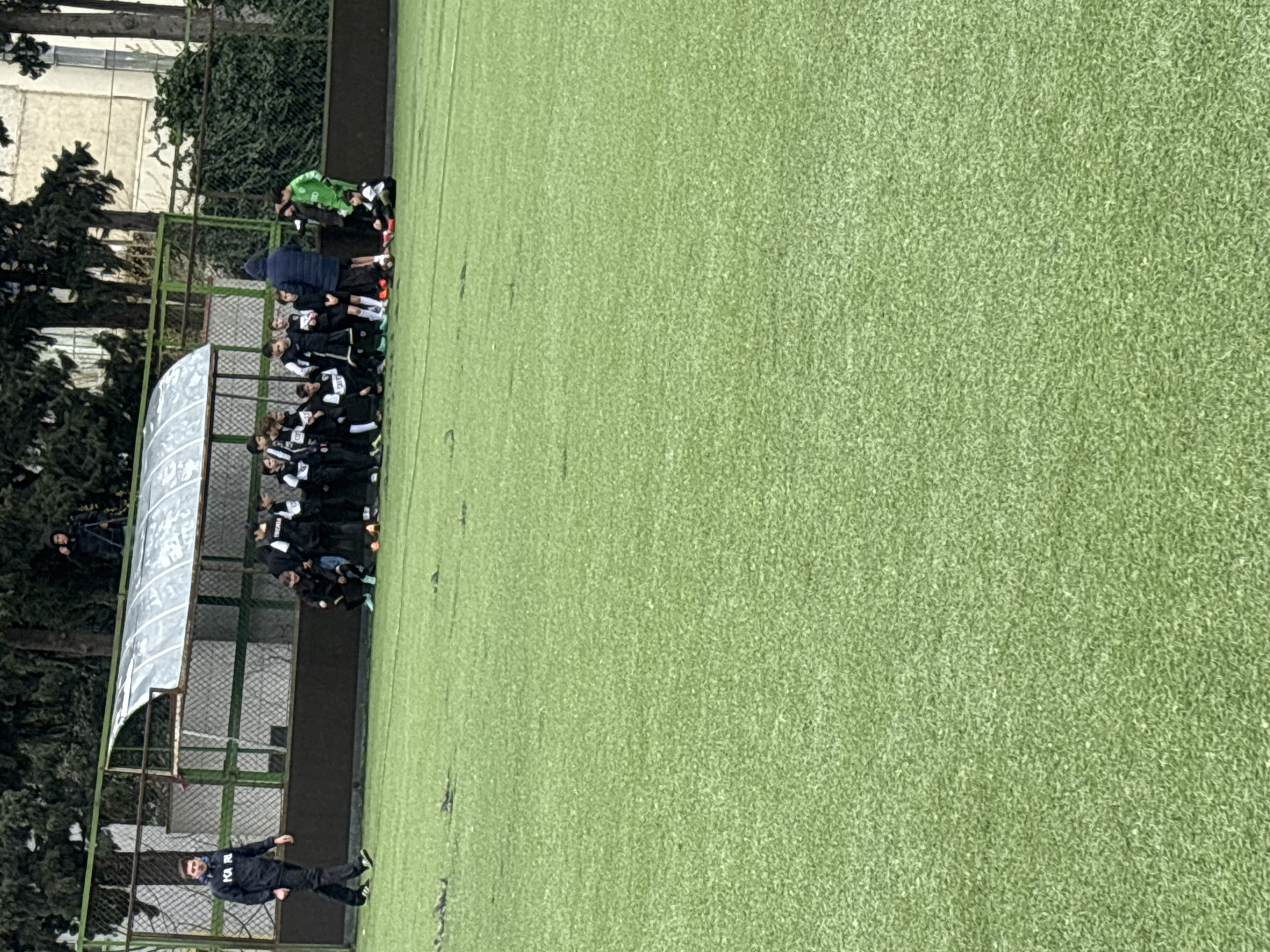 A soccer team, presumably youth, stands near a shelter on the edge of a soccer field in Tbilisi, Georgia. The field is covered in artificial green turf. A man in black, possibly a coach, walks along the field’s edge, away from the team. Trees are visible along the periphery, and a building stands in the background.