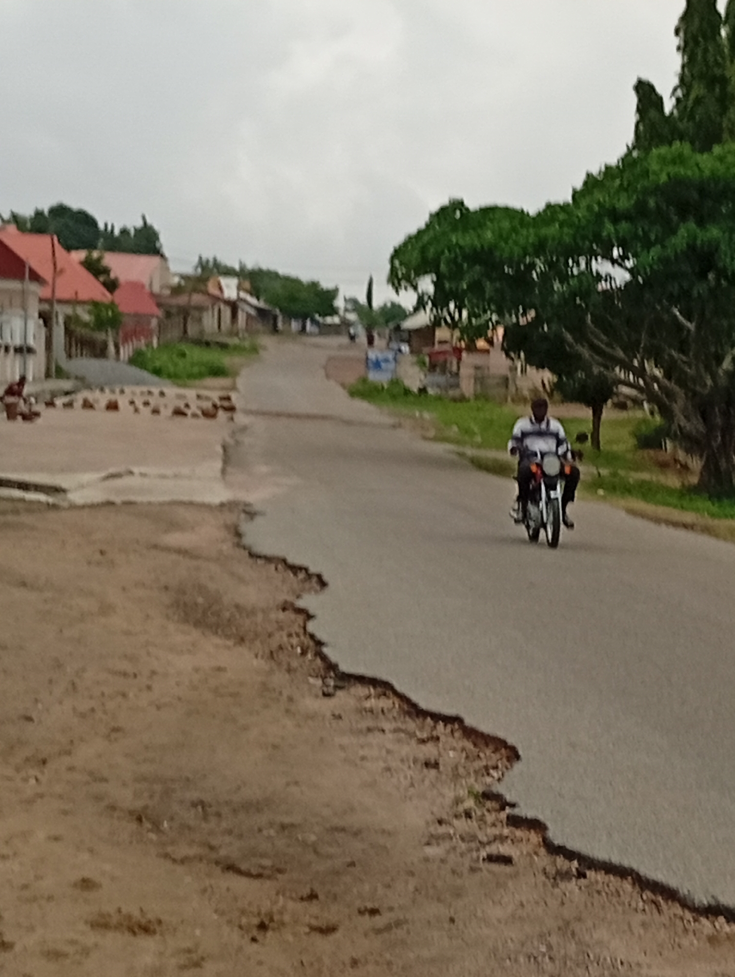 The image shows a street scene in Lokoja, Nigeria. A black man is riding a motorcycle toward the camera. A second person is on the back of the motorcycle. To the left of the road is a dirt area. A concrete sidewalk is adjacent to the road, with bricks scattered on it. Buildings with red roofs are visible in the distance. Trees line the right side of the road. The sky is overcast. The road surface shows signs of disrepair.