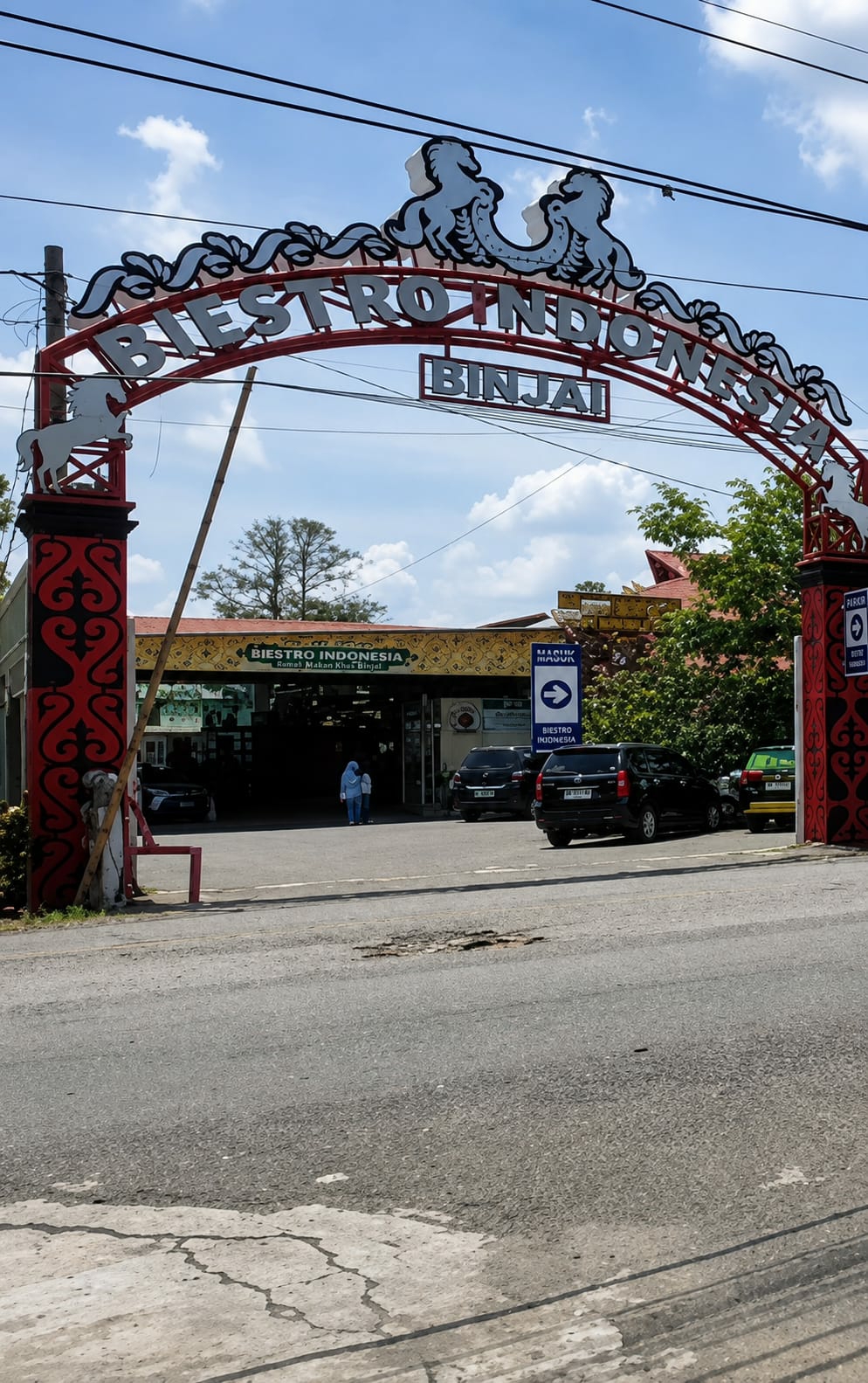 The image shows the entrance archway to "BIISTRO INDONESIA BINJAI" in the city of Binjai, Indonesia. The arch is red with white decorative elements depicting horses and swirls. Below the arch, a sign reads "BIISTRO INDONESIA" with smaller text underneath, "Rumah Makan Khas Binjai" which translates to "Binjai Specialty Food House." Several cars are parked in the lot in front of the building, and two people, one wearing a light blue headscarf, are walking towards the entrance. The scene is set outdoors with a blue sky and white clouds above. The road in the foreground appears to be asphalt with some visible cracks and damage. The building itself has a yellow facade with intricate dark red and black patterns on the pillars. A blue directional sign points towards "MASUK" (Entrance) and "BIISTRO INDONESIA."