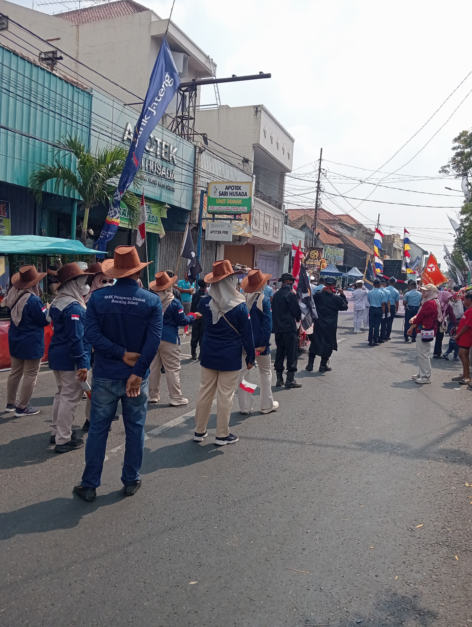A street-level, outdoor, full-shot image depicts a parade or procession taking place on a road in the city of Bintoro, Indonesia. The scene is dominated by a large group of individuals, many of whom are wearing matching navy blue shirts with "SMK Pelayaran Demak Boarding School" printed on them. They are also wearing brown, wide-brimmed hats and tan pants. Several people in this group are holding flags, some of which are red and white, possibly representing the Indonesian flag.
To the left of the frame, buildings line the street, including one with a blue facade and signage that includes "APOTEK" and "HUSADA." A palm tree is visible near this building. Several other flags, some with colorful designs, are planted along the street, adding to the festive or ceremonial atmosphere.
In the background, more people are visible, including some in uniforms, and additional flags are displayed. The road surface is asphalt, and shadows are cast on the ground, indicating a bright, sunny day. The overall impression is that of an organized event involving students or a school group in a public space.