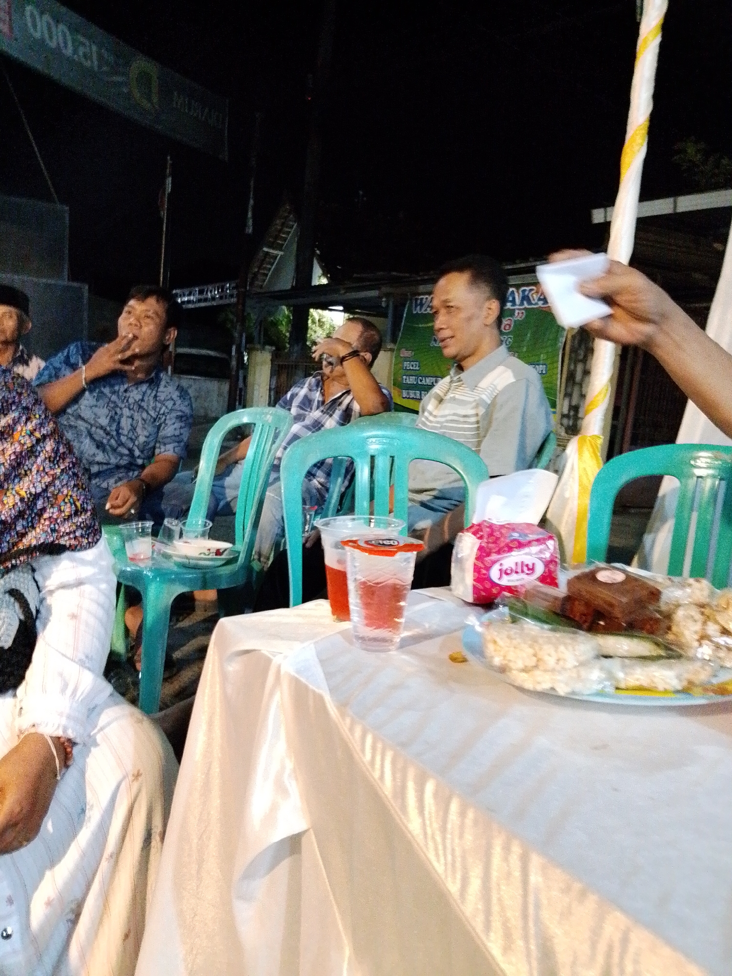 The image depicts an outdoor gathering at night, likely a social event, possibly in Trangkil, Indonesia. In the foreground, a white tablecloth covers a table laden with snacks. Among the snacks are packets of what appear to be puffed rice or crackers, a small brown rectangular cake, and a pink tissue box with the brand "Jolly" visible. Two glasses with red liquid are also on the table.
Behind the table, several individuals are seated on green plastic chairs. On the left, a man in a blue patterned shirt is smoking a cigarette. Next to him, a man in a plaid shirt is also seated. Further to the right, a man in a light grey and white striped shirt is looking towards the right. Another person, partially visible on the far left, is wearing a white shirt with a subtle pattern.
In the background, there's a dark setting with some illuminated signs and structures. One sign in the upper left corner displays "000.21". Another sign with green lettering is partially visible behind the seated individuals, with words like "TIS CAMPU" and "LUBUR" discernible. A tall, striped yellow and white pole is prominent on the right side of the image. The overall atmosphere suggests a casual, communal event taking place after dark.