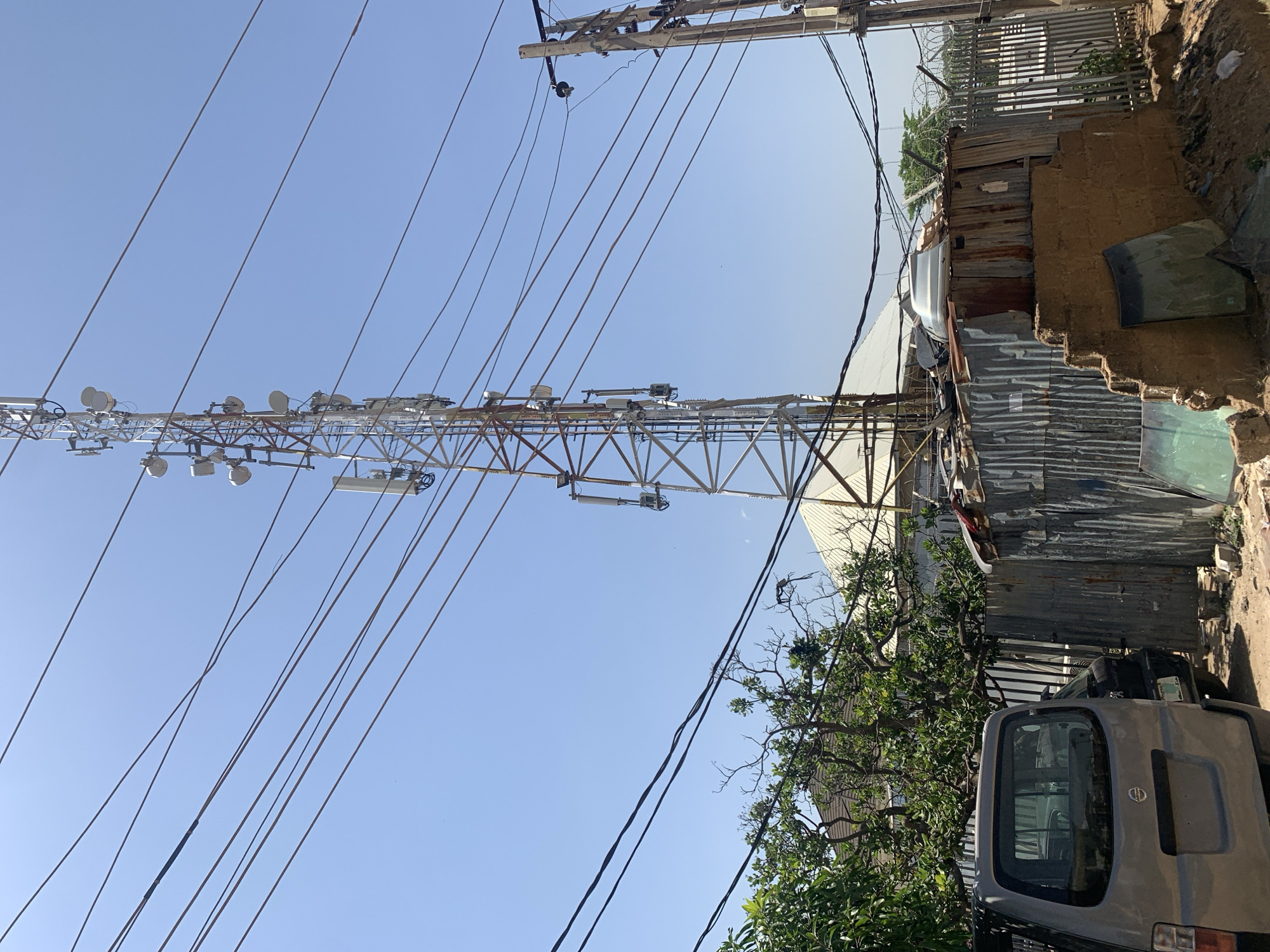 The image depicts an outdoor scene in Rafin Par, Nigeria. A tall telecommunications tower, fitted with multiple antennas and electrical cables, dominates the left side of the frame against a clear blue sky. Adjacent to the tower is a makeshift structure constructed of corrugated metal sheets and what appears to be earthen materials. A gray van is parked near the structure, partially obscured by foliage. Overhead, power lines are strung across the frame, converging toward a utility pole. The overall environment suggests a mixed urban/residential area with possibly informal construction.