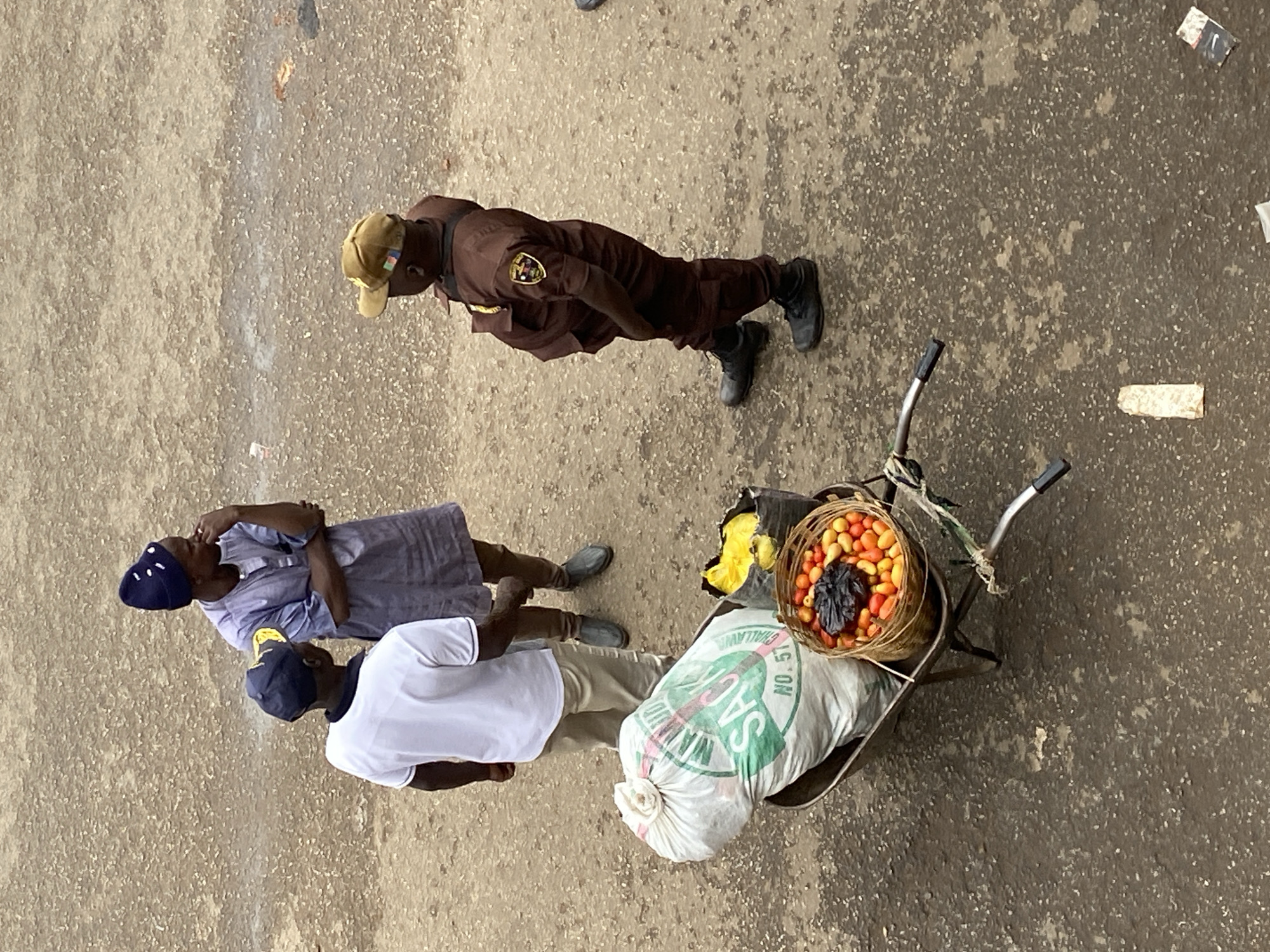 The image, taken from an elevated perspective, depicts a scene in Bukuru, Nigeria. Three individuals are visible: two adults and a uniformed officer. The officer, wearing a brown uniform with a patch on the upper arm and a tan cap, stands slightly apart. The adults, one wearing a white shirt and tan pants with a blue cap, the other in a blue head covering and a light blue dress, stand closer together. A wheelbarrow loaded with a large sack and a basket containing tomatoes is positioned near them. The surrounding environment appears to be a paved street or open area.