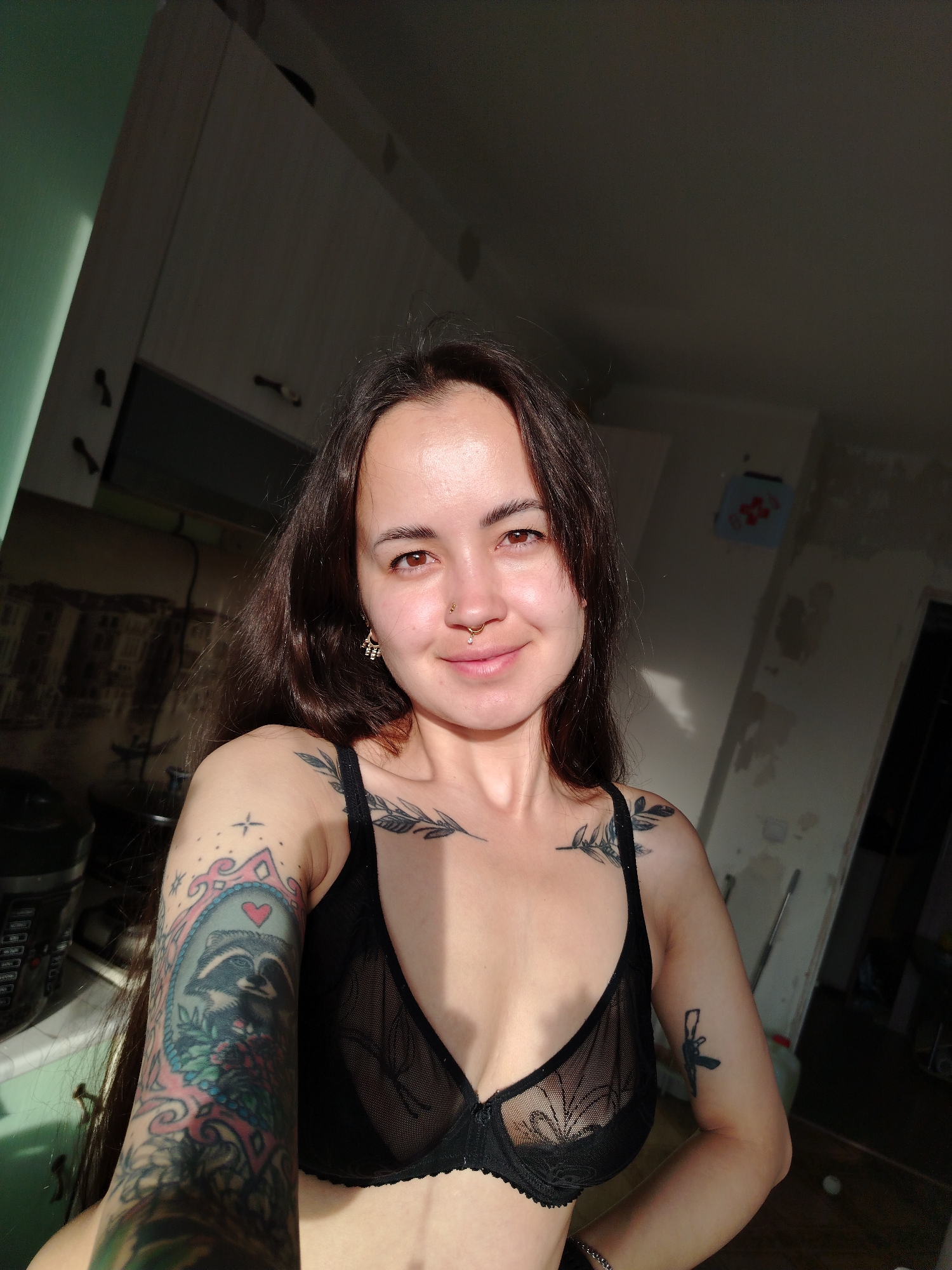 The image features a young woman with long dark hair, light brown eyes, and fair skin, smiling gently at the camera. She has a nose ring with a small gem, a septum piercing, and an ornate earring on her right ear. She is wearing a sheer black lace bralette.
She displays several tattoos: a prominent colorful full sleeve on her left arm featuring a raccoon in a decorative frame with a heart, flowers, and stars. On her shoulders and chest, there are tattoos of leafy branches. Another tattoo of a firearm is visible on her right upper arm.
The woman appears to be taking a selfie in an indoor setting, likely a kitchen or utility room, which seems to be undergoing renovation or is unfinished. The background shows partially stripped walls with exposed plaster and remnants of wallpaper, as well as white kitchen cabinets on the left. A section of the wall on the left is painted light green. A small blue box with a red cross or design is affixed to one of the walls. Strong sunlight illuminates her face and parts of the room, suggesting a daytime setting. The woman appears relaxed and content. No discernible text is visible. The location context is Noviy, Russia.