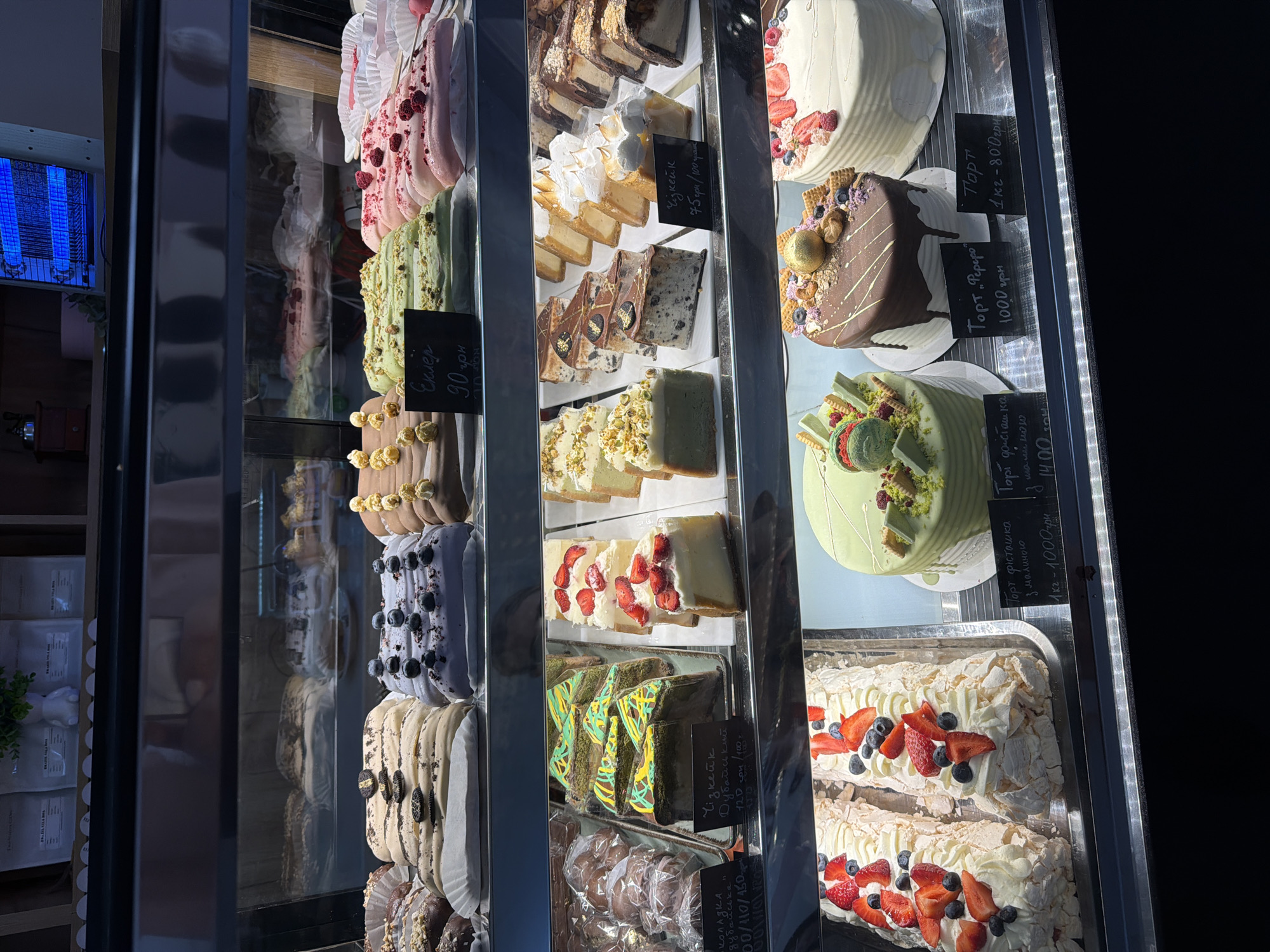 This is a full-frame, eye-level, medium shot of a display case filled with various desserts. The desserts are arranged on multiple shelves and include cakes, pastries, and possibly ice cream.
The display case is illuminated, highlighting the colorful and ornate nature of the desserts. On the top shelf, there are slices of cheesecake topped with whipped cream and berries, and a whole white cake adorned with strawberries and blueberries. Below these, a chocolate cake with a drip glaze, nuts, and berries is visible. Further down, there are pastel-colored pastries, some appearing to be eclairs or similar filled items, decorated with blueberries and other toppings. There are also slices of cheesecake with fruit and layered cakes with green and yellow frosting. Towards the bottom, two large meringue rolls topped with strawberries and blueberries are prominently displayed.
Several black price tags with white handwritten text are placed in front of some of the desserts, indicating prices in "грн" (hryvnia) and "гри" (hryvnia), with some also mentioning weight in kilograms. Text visible includes "Чізкейк", "Еклер", "Торт фісташка", "Торт „Фереро", and "КОЛАДКА". The overall scene suggests a bakery or dessert shop. The lighting suggests it could be daytime, or the interior is well-lit. There are no people visible in the image.