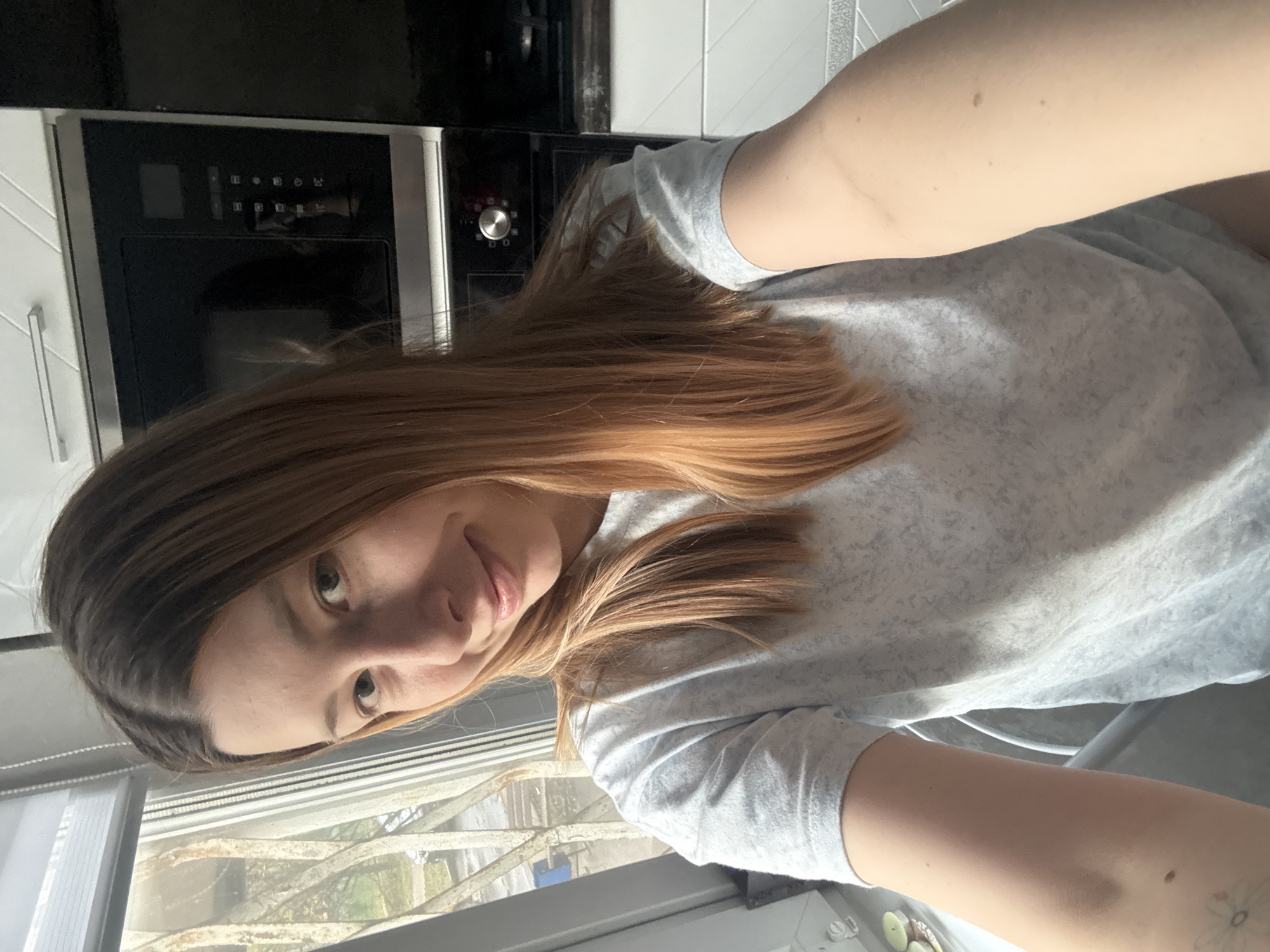 A selfie photograph, originally taken in portrait orientation but displayed rotated 90 degrees clockwise, captures a young woman indoors. She has long, light brown to auburn hair, light eyes, and visible freckles across her face. The woman is wearing a light grey t-shirt with a subtle speckled pattern and looks directly at the camera with a calm, slight smile.
The indoor background features a modern black kitchen appliance, likely an oven or microwave, with a digital display panel and a silver control knob. White tiled walls are visible behind the appliance. Behind the woman, a window offers a view of the outdoors. Bare tree branches, possibly frosted or covered in snow, are prominent, suggesting cold weather or a winter season. Patches of what appears to be snow or ice are visible on the ground or a distant surface outside. The lighting indicates it is daytime. While symbols are visible on the appliance's control panel (e.g., power, fan, grill), no clear alphanumeric text is discernible. The scene is contextualized as being in Moscow, Russia.
