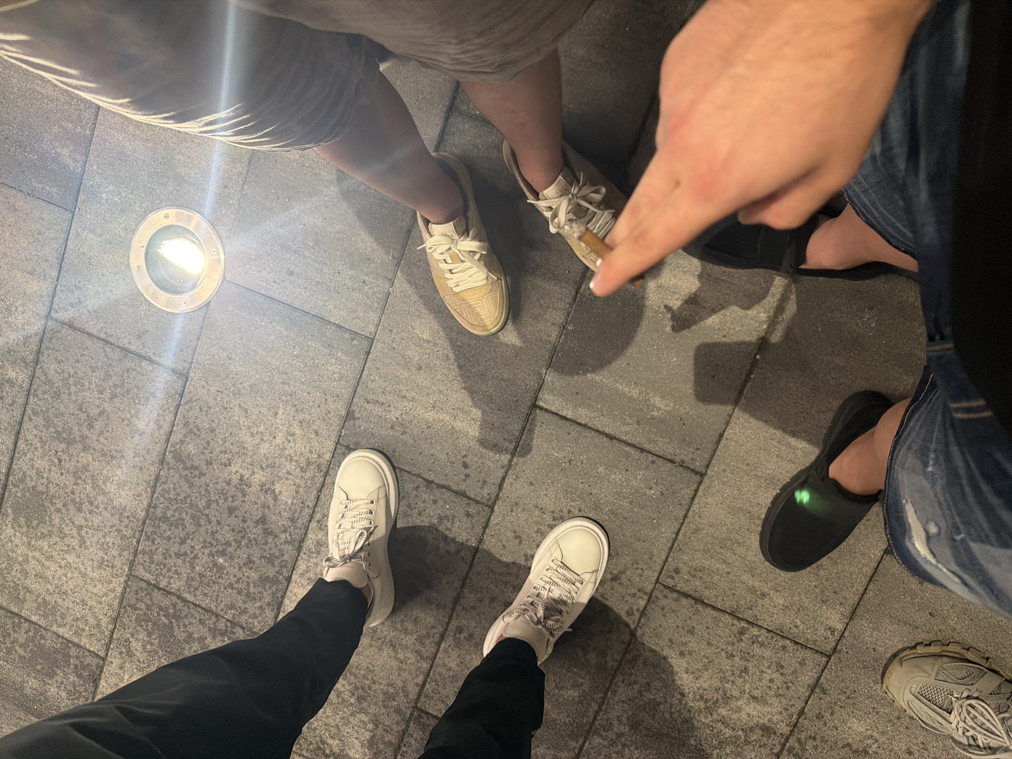 The image, taken from an overhead perspective, captures the lower bodies and feet of at least four individuals standing on a paved outdoor surface in Sofia, Bulgaria, likely during the evening or night. The ground is composed of grey, rectangular stone or concrete slabs.
In the upper left, a person wears light-colored, low-top sneakers with a subtle patterned design on the sides and light-colored shorts. A bright, circular in-ground light fixture is positioned nearby, casting strong light and reflections onto the surroundings.
To the right, a hand is visible holding what appears to be a lit cigar or cigarette. This person wears a pair of light-colored, athletic-style sneakers with prominent laces, similar in color to the first pair. Their lower legs are also visible.
Towards the bottom center, a person is wearing off-white, thick-soled sneakers with patterned laces, paired with black trousers.
On the right side of the frame, another individual wears black slip-on shoes, one of which features a faint glowing green emblem on the top. This person is dressed in blue jeans. Below their foot, a grey, chunky sneaker with intricate lacing is partially visible, indicating a fourth person.
The strong illumination from the ground light fixture and the overall dimness of the surroundings suggest the scene takes place after dusk. No discernible text is visible on clothing or objects. The individuals appear to be casually gathered outdoors.
