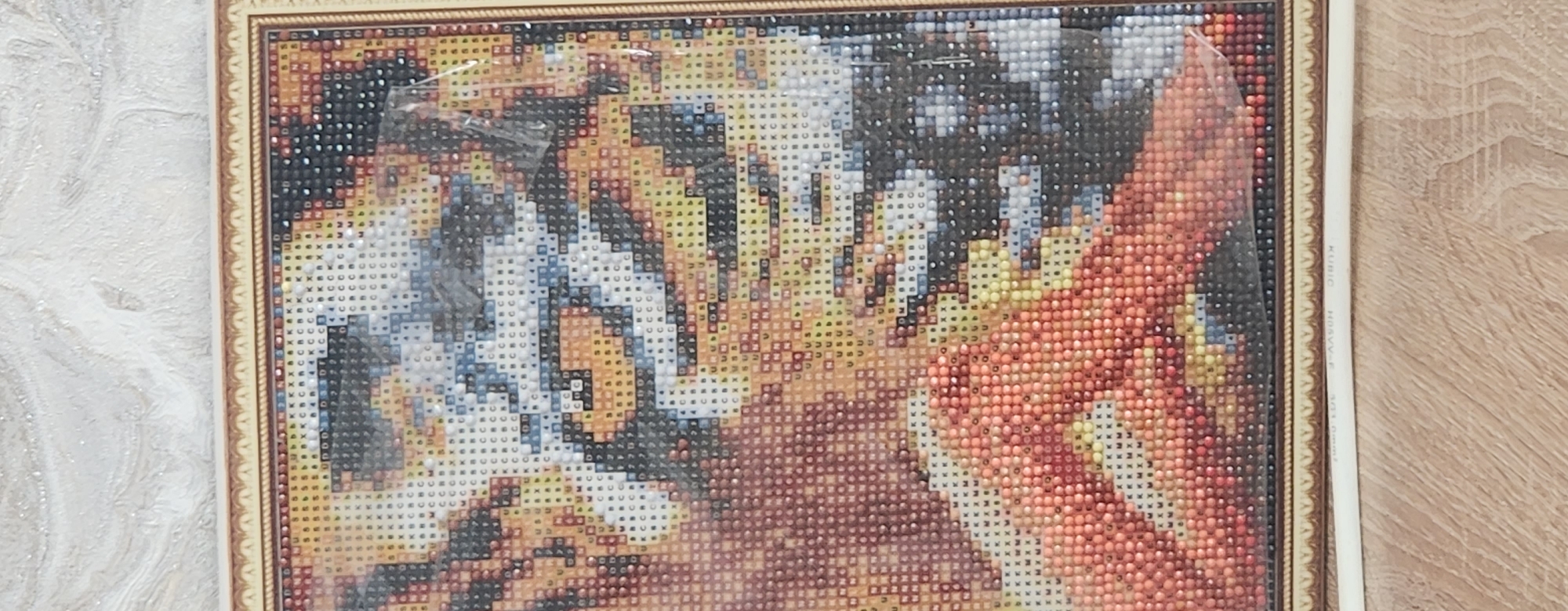 The image displays a close-up of a diamond painting, likely a tiger's face, framed in a metallic border. The painting is composed of tiny, colored square or diamond-shaped beads, arranged in a grid to form the image. The beads are visible in shades of orange, black, white, yellow, and blue, suggesting the intricate detail of the artwork. The painting is covered with a protective plastic sheet, which is slightly creased in places. The background behind the painting is a textured, pale wall. Below the painting, a wooden surface is visible, and a white cable with markings that include "KUBIC" and "301.0mm²" rests on the wood. There are no people or explicit actions depicted in this image. The scene does not provide any visual cues to indicate it is located in Kazan, Russia.
