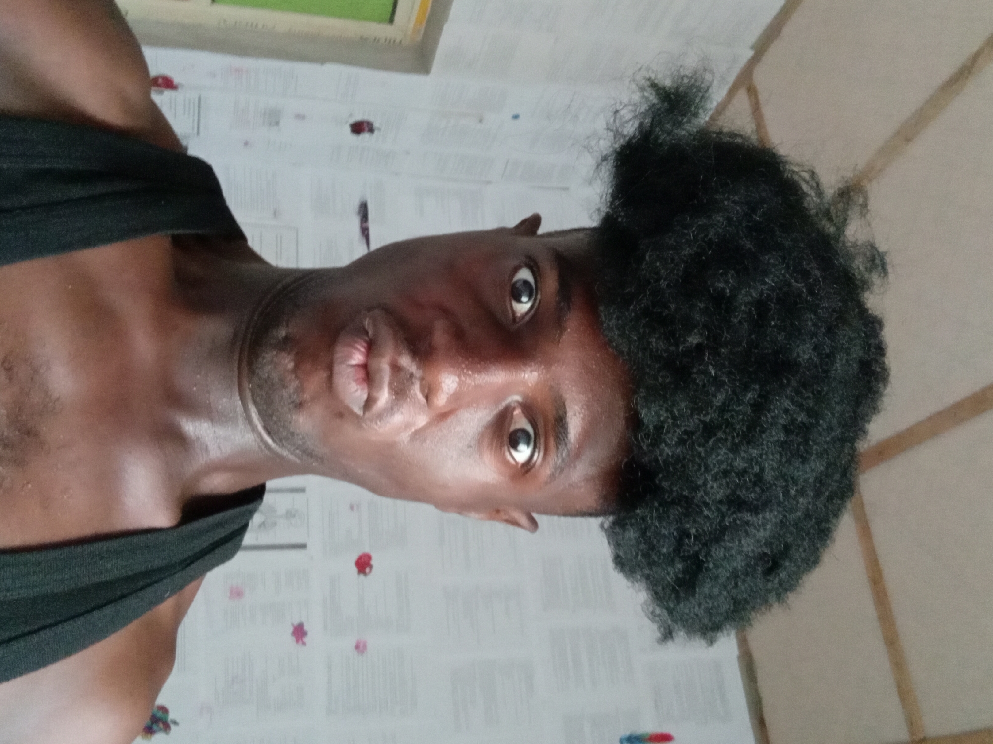 A man with dark skin and black, curly hair is pictured close-up, facing the camera with pursed lips. He is wearing a black tank top. The background appears to be a room with printed papers covering the wall and wooden paneling. The location is unspecified in Akwete, Nigeria.