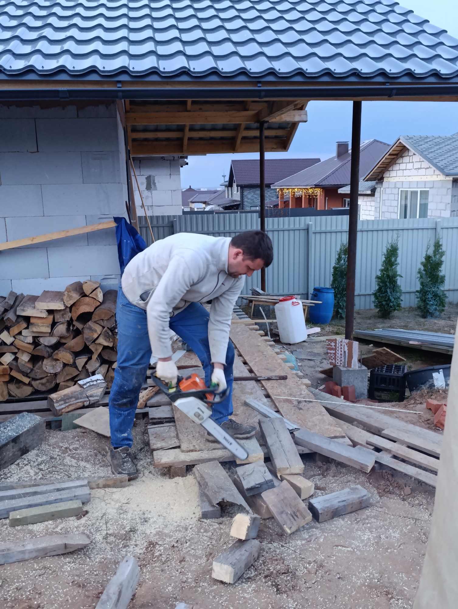 A man is operating a chainsaw outdoors, cutting wooden planks amidst sawdust and wood debris. He is wearing a white sweatshirt, blue jeans, and gloves. Behind him, there's a stack of firewood against a building under construction with a grey metal roof. In the background, houses are visible beyond a fence. The setting appears to be a residential area, possibly in the city of Chaikovsky, Russia, based on the architectural style of the houses and fences. Other objects present include blue barrels, a white container, bricks, and greenery.