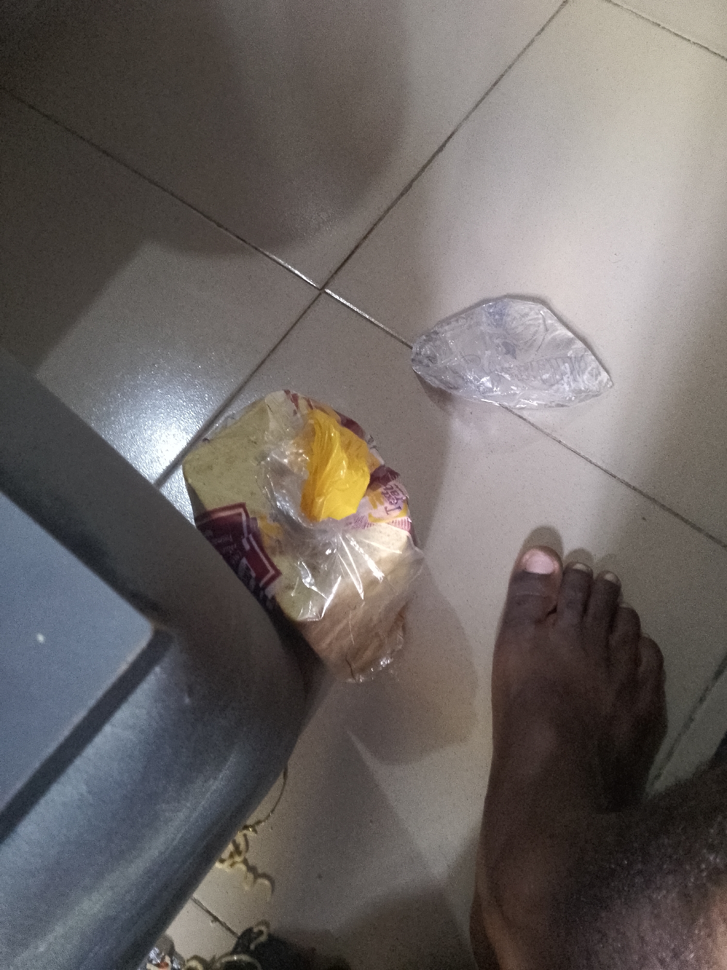 An overhead, medium shot shows a foot of a dark-skinned person on a tiled floor. The person is barefoot and their toes are visible. To the left of the foot, a loaf of sliced bread is partially wrapped in clear plastic. Above and to the left of the bread, a crumpled piece of clear plastic is visible. To the left of the bread, a portion of a gray object, possibly a piece of furniture, is also visible. The floor tiles are light gray and appear to be in a domestic setting. The lighting suggests an indoor environment, possibly a kitchen or hallway. The scene is static, with no apparent action or interaction occurring.