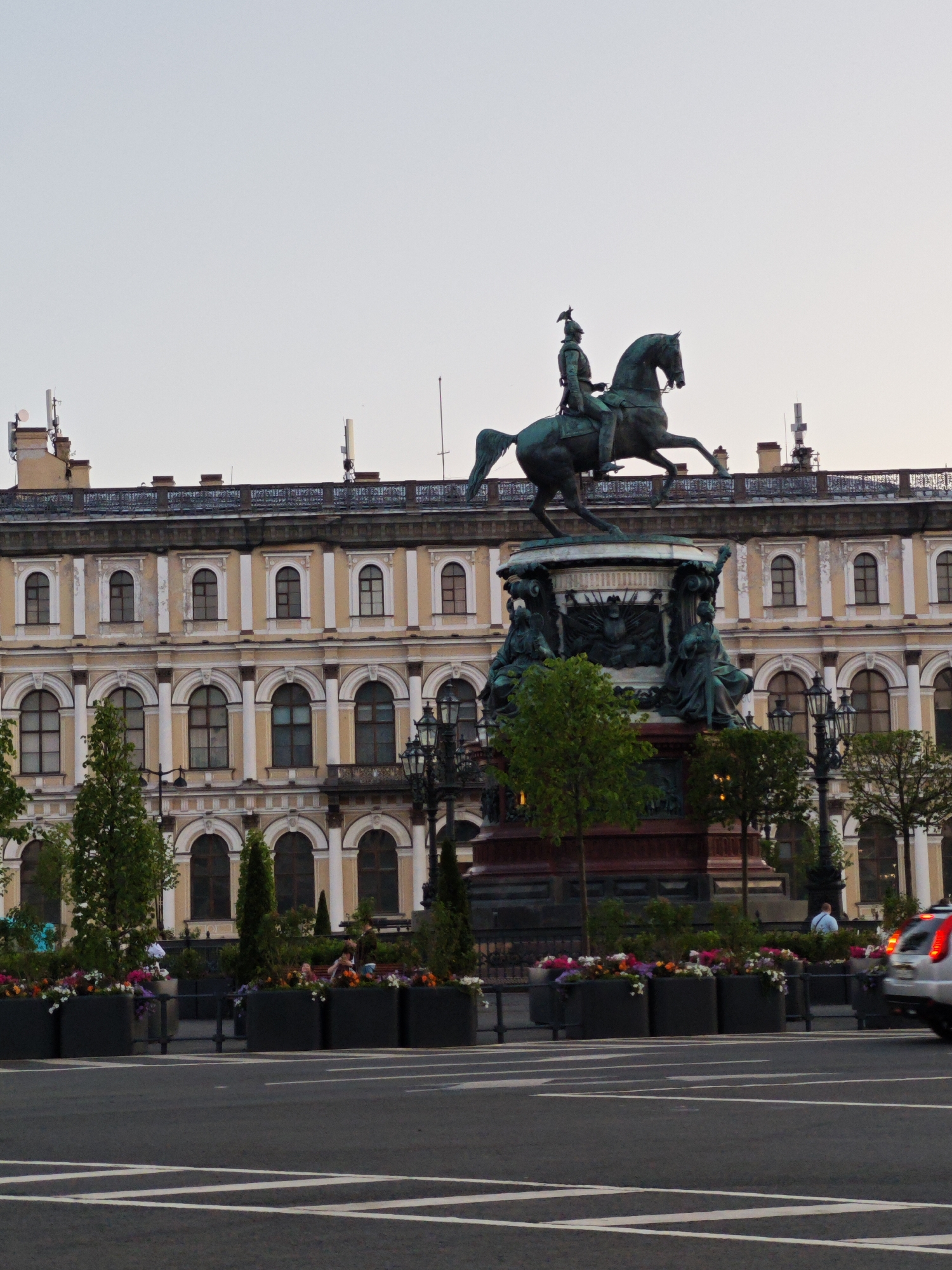 This image captures the equestrian statue of Nicholas I in Saint Isaac's Square, Saint Petersburg, Russia. The monumental bronze statue features Emperor Nicholas I on a rearing horse, facing slightly left. The horse balances on its hind legs. The emperor wears military attire, including a helmet with a plume. The statue rests on a dark, multi-tiered pedestal adorned with four allegorical female figures at its base, each seated and facing outward.
Behind the statue stands a large, multi-story building. Its facade is a light yellow-ochre color, featuring numerous arched windows with dark glass. White architectural elements, including pilasters and decorative window surrounds, accent the building's horizontal and vertical lines. The roofline includes a dark, ornate railing, several chimneys, and multiple modern cellular antennae or communication towers.
In the foreground, a dark asphalt road with white diagonal and straight lane markings occupies the bottom third of the frame. Separating the road from the monument and building is a planted area. This area contains large, dark rectangular planters filled with a variety of colorful flowers, including shades of pink, purple, orange, and white. Several green trees and bushes of varying heights are interspersed among the planters and around the base of the monument. Two ornate black lampposts with multiple luminaires are visible on the right side.
A few people are present at street level. On the right, behind the planters, a person wearing a light blue shirt is visible from the back, standing. Further left and partially obscured by trees and planters, two or three individuals appear to be seated on a bench. On the far right, a silver car is partially visible, moving away from the viewer, with its red taillights illuminated. The sky above is a pale, uniform grey, suggesting an overcast day or twilight conditions.