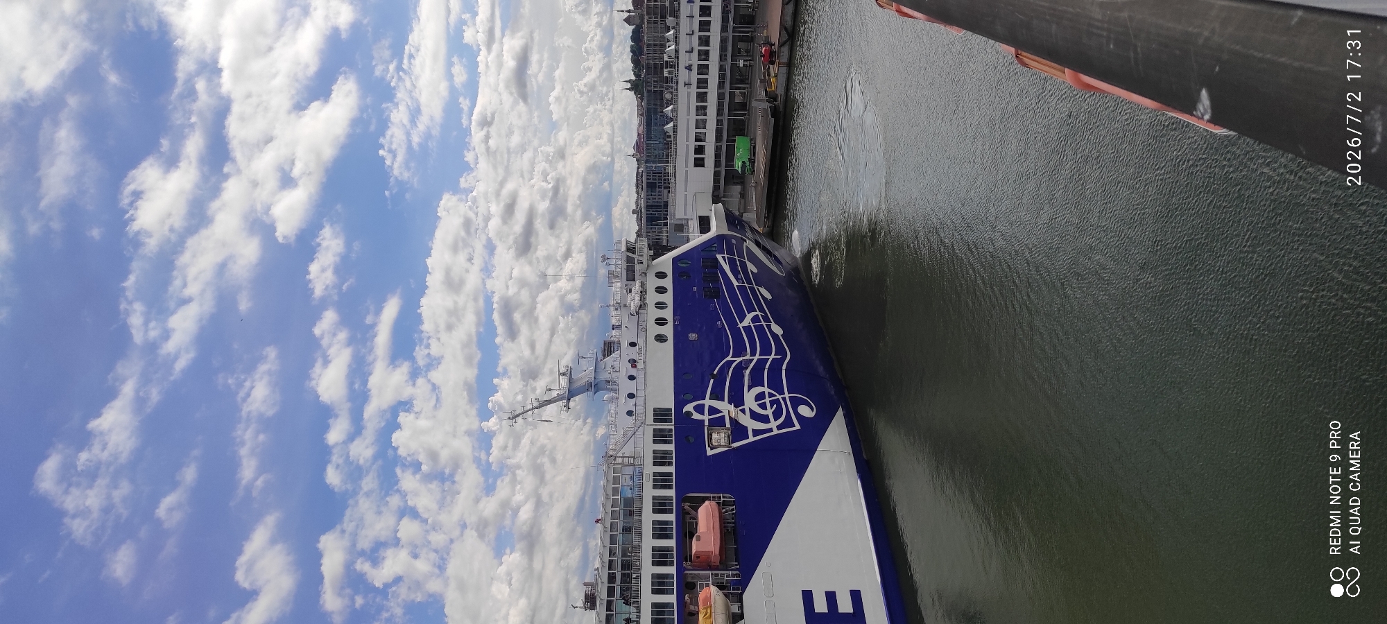 This is a media file featuring a ferry docked in Tallinn, Estonia. The ferry is primarily blue and white, with musical notes painted on its side. The sky is filled with white, puffy clouds against a bright blue backdrop, suggesting fair weather. The water next to the ferry is calm and dark green. In the background, buildings that appear to be part of the Tallinn cityscape are visible. The image is taken from an elevated perspective, likely from another ship or a tall structure.
The time of day is indicated by the text "2026/7/2 17:31", suggesting late afternoon. The text "REDMI NOTE 9 PRO AI QUAD CAMERA" is also visible in the lower right corner of the image, indicating the device used to capture the photo. There are no people visible in the frame, but the ferry's presence suggests it is either preparing for departure or has recently arrived. The scene conveys a sense of calm and travel.