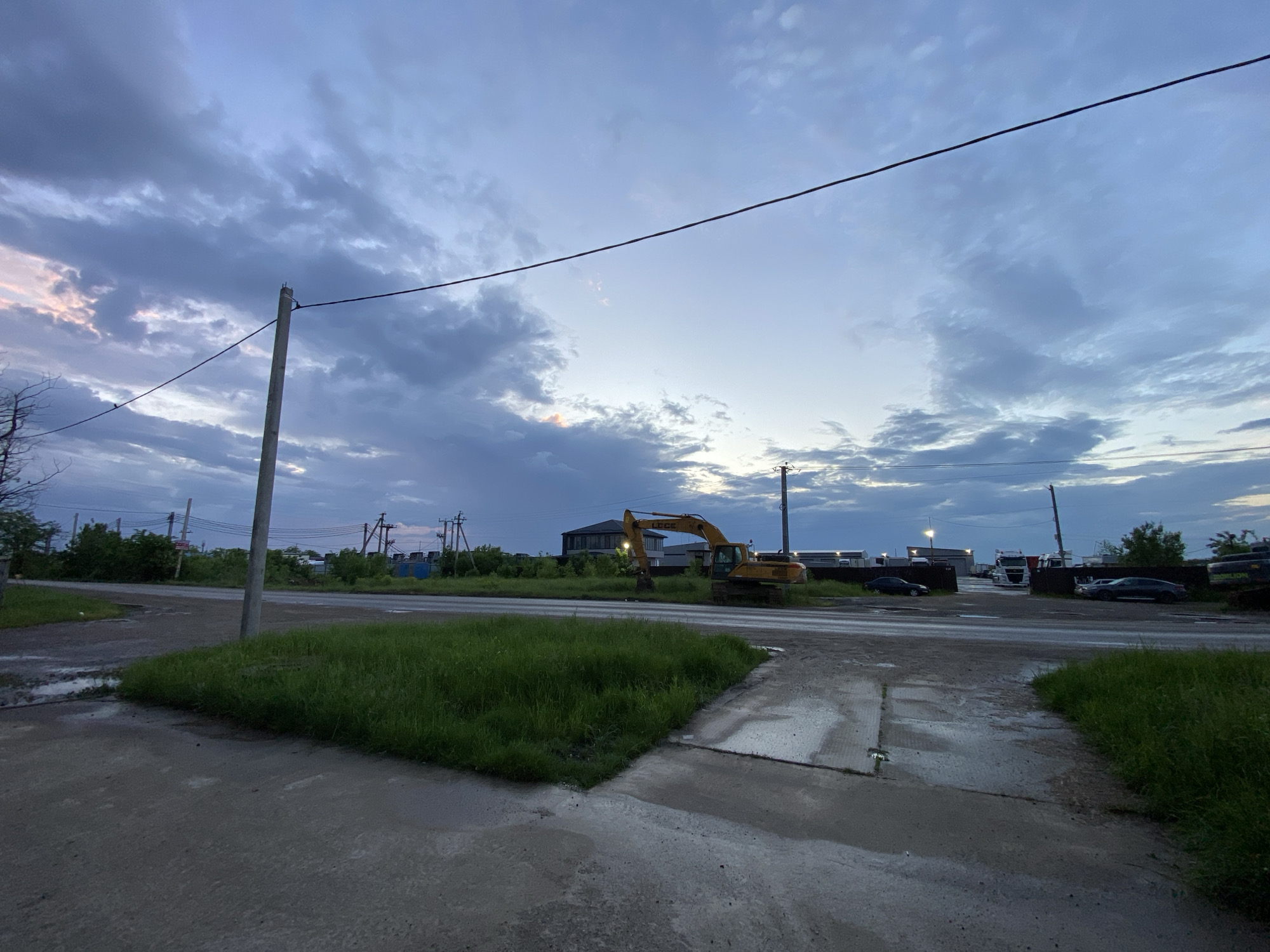 The image depicts an outdoor scene in the city of Neftyanik, Russia. A concrete road and adjacent grassy patches dominate the foreground, showing signs of recent rain, with puddles visible. A utility pole stands on the left edge of the road, power lines stretch across the sky. In the middle ground, an excavator is parked. In the background, there are several buildings, vehicles (cars and trucks), and more vegetation. The sky is cloudy.
