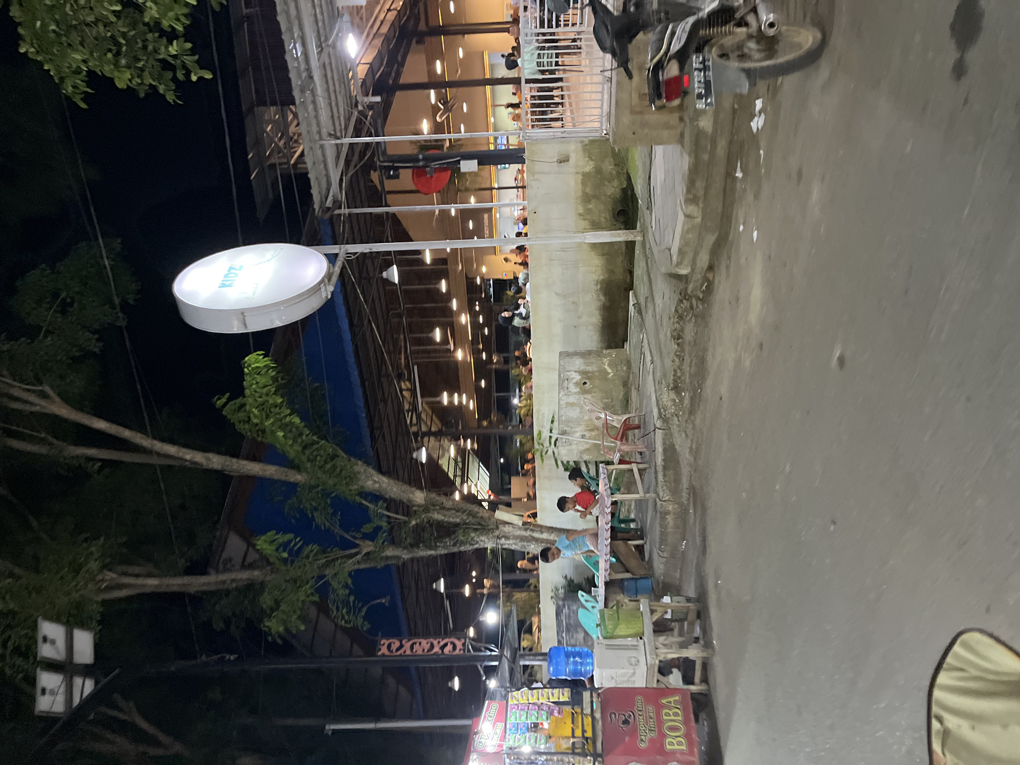 The image depicts a street scene at night, likely in Langsa, Indonesia. The setting is an outdoor dining area or a market, with a building in the background illuminated by numerous lights. In the foreground, a paved street is visible.
Several people are present, some seated at tables and others standing. There are also objects such as chairs, tables, a water dispenser, a cooler, and a motorcycle. A prominent circular illuminated sign with the word "KIDZ" in light blue text hangs from a pole. Further down the street, a stall with various items for sale, including drinks and packaged goods, is visible. A large sign on this stall reads "Cappuccino" and "BOBA".
The time of day is clearly night, indicated by the darkness and the artificial lighting. The weather appears to be clear, as there are no signs of rain. The overall atmosphere suggests a lively evening, with people dining and shopping. The presence of the "BOBA" and "Cappuccino" signs, along with the general setup, hints at a food or beverage establishment. The location context of Langsa, Indonesia, provides a geographical anchor for the scene.