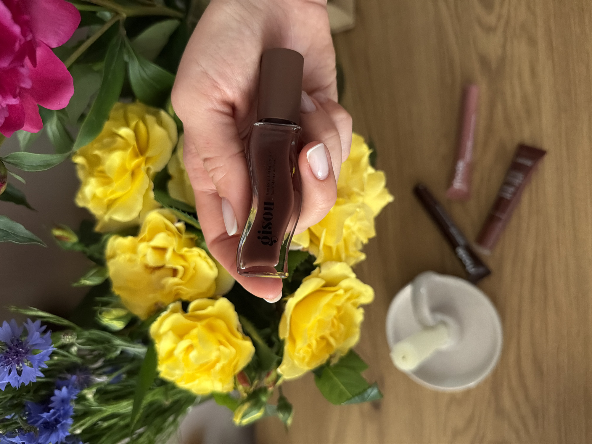 The image is a top-down shot featuring a hand holding a small bottle of Gisou honey-infused lip oil. The bottle has a brown cap and a clear base revealing a brownish-red oil. The hand has a French manicure.
The background includes a bouquet of flowers with yellow roses, pink peonies, and blue cornflowers. To the right of the bouquet are a few makeup products: a pink tube, a brown tube, and a dark brown eyeliner. There is also a small, white mortar and pestle. The surface the items are resting on appears to be a light brown wooden table. The location appears to be in a personal residence.