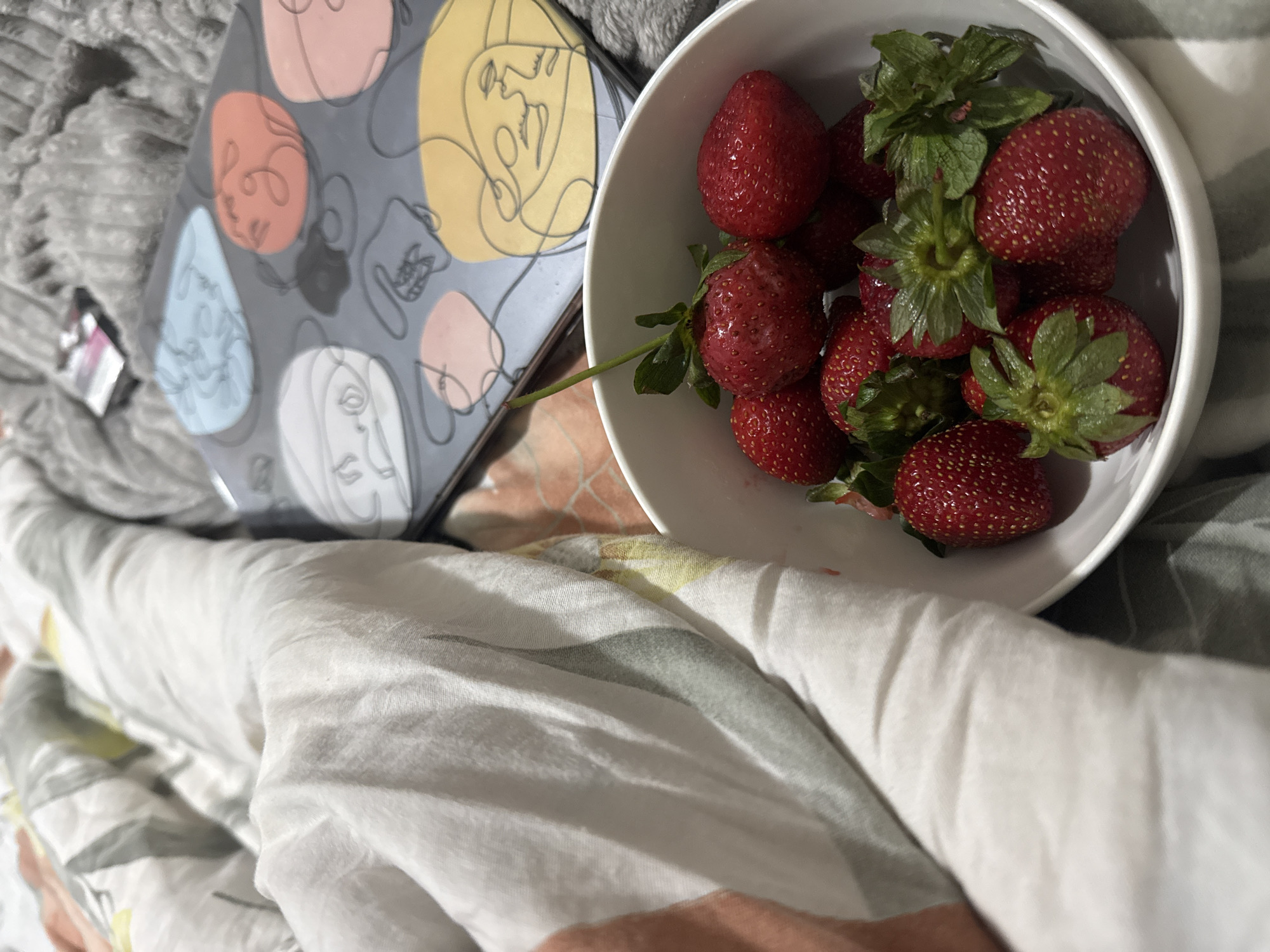 A white bowl containing strawberries is placed on a bed covered with white bedding and a blanket. Next to the bowl is a gray object with colorful face designs. The location is implied to be Batumi, Georgia, but there are no visual clues to confirm this. There are no people visible.