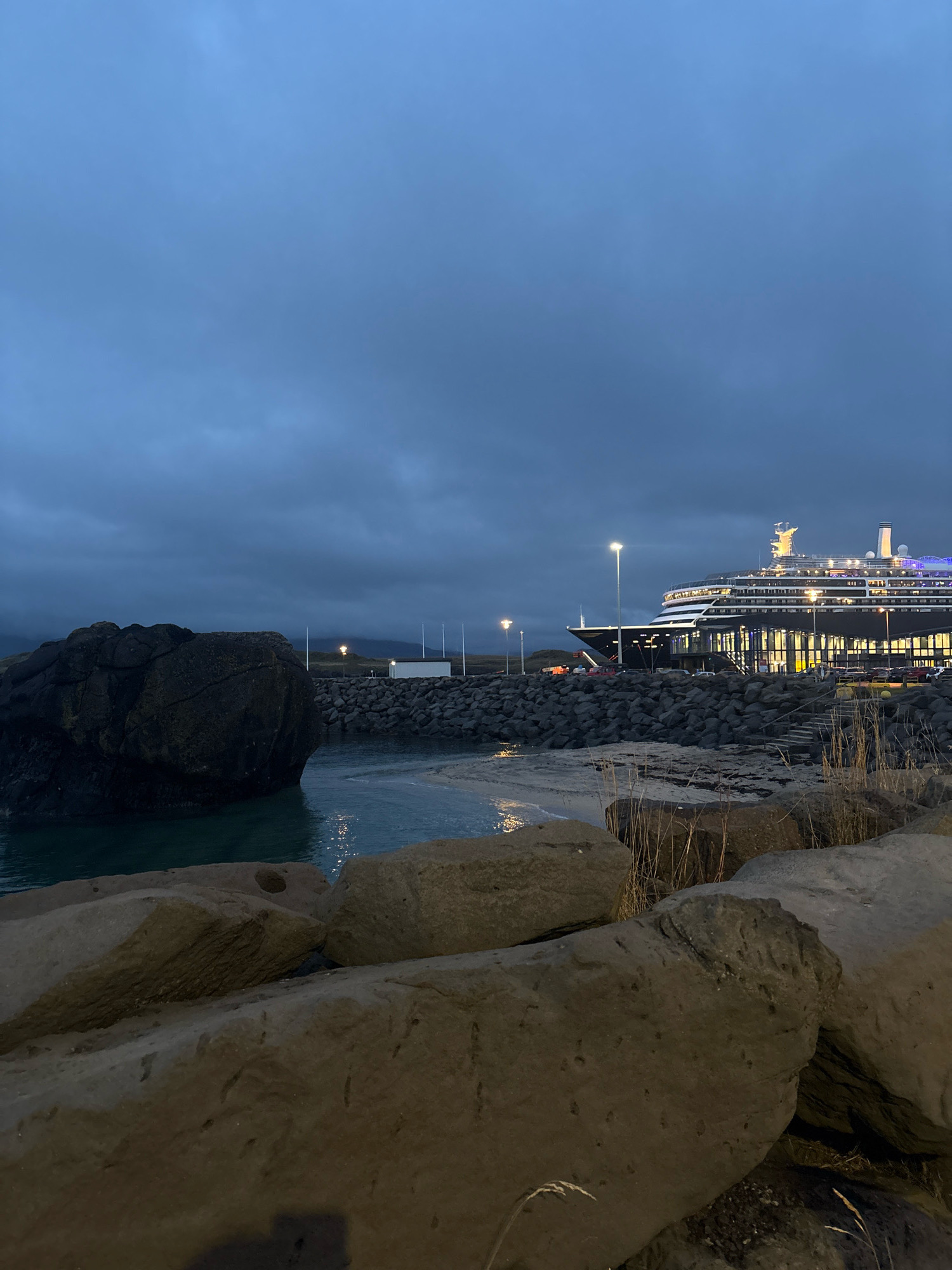 This image depicts a twilight scene at the Grandi marina area in Reykjavik, Iceland. In the foreground, large, rough-hewn rocks line the shore. To the left, a substantial dark boulder juts into the calm, turquoise water. Beyond this, a small sandy cove is visible. Further out, a large cruise ship is docked at a pier, its lights illuminating the dark hull and the terminal building. The terminal has a modern, glass-fronted facade, and the area around it appears to be a parking lot with several vehicles. A streetlamp casts a pool of light on the pier. The sky is overcast and dark blue, suggesting the late hour or impending weather. In the middle ground, a white rectangular building with a gabled roof stands near the water's edge, and several flagpoles are visible behind it. Dry grass and reeds grow along the rocky shore in the mid-ground and foreground. The overall impression is one of a quiet, functional harbor at dusk.