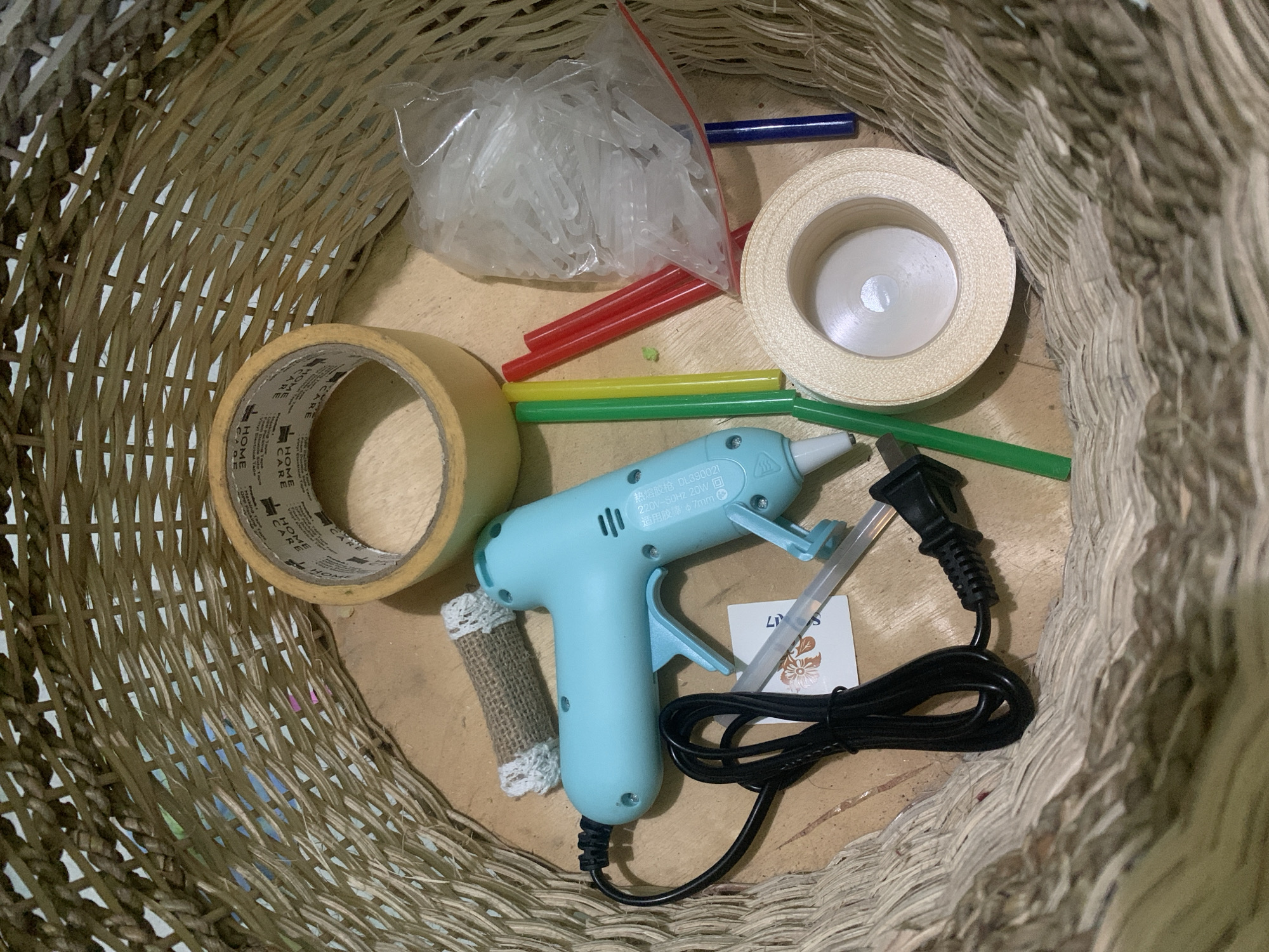 In a woven basket, a turquoise hot glue gun with a black cord is positioned next to several sticks of glue, some yellow, red, and blue. A roll of beige ribbon lies to the right, and a plastic bag filled with clear plastic fasteners is also present. To the left, a roll of yellow tape is partially visible, and a small label with floral imagery is near the glue gun. The basket itself is made of natural, light-colored fibers. The scene is set on a wooden surface, likely a table or floor, with the natural fibers of the basket spilling over the edges. This arrangement suggests a crafting or DIY project in progress or planned, but there are no people or actions depicted, nor any specific indicators of the city of Kuala Pembuang, Indonesia.