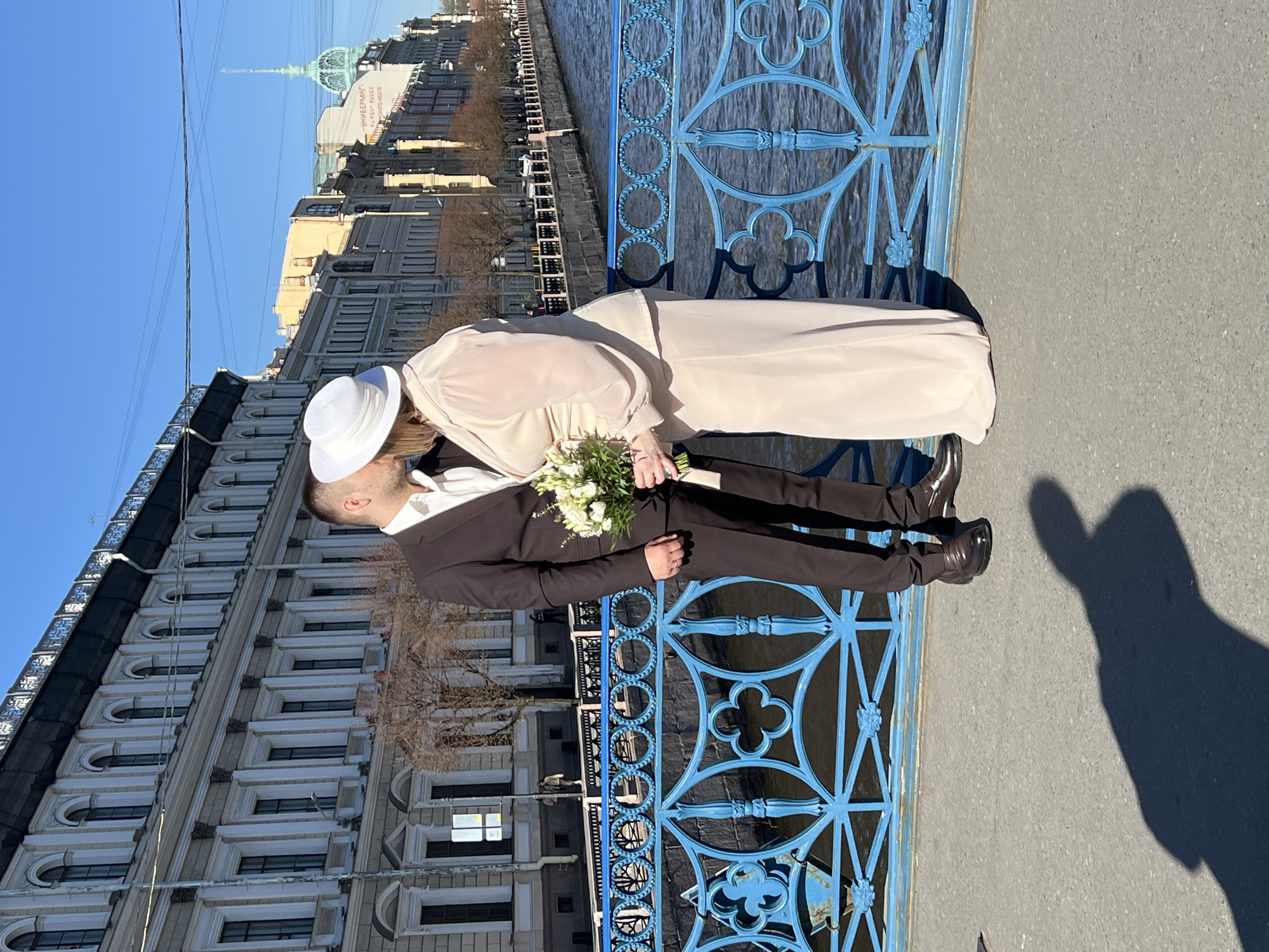 A couple in Saint Petersburg, Russia are kissing on a bridge. The man is wearing a brown suit, and the woman is wearing a long, light pink dress and a white hat. She is holding a bouquet of white flowers. They are standing on a blue bridge, with a river visible beneath the bridge. In the background is a building with a green dome. The sky is clear and blue. A long shadow is cast on the right side of the image.