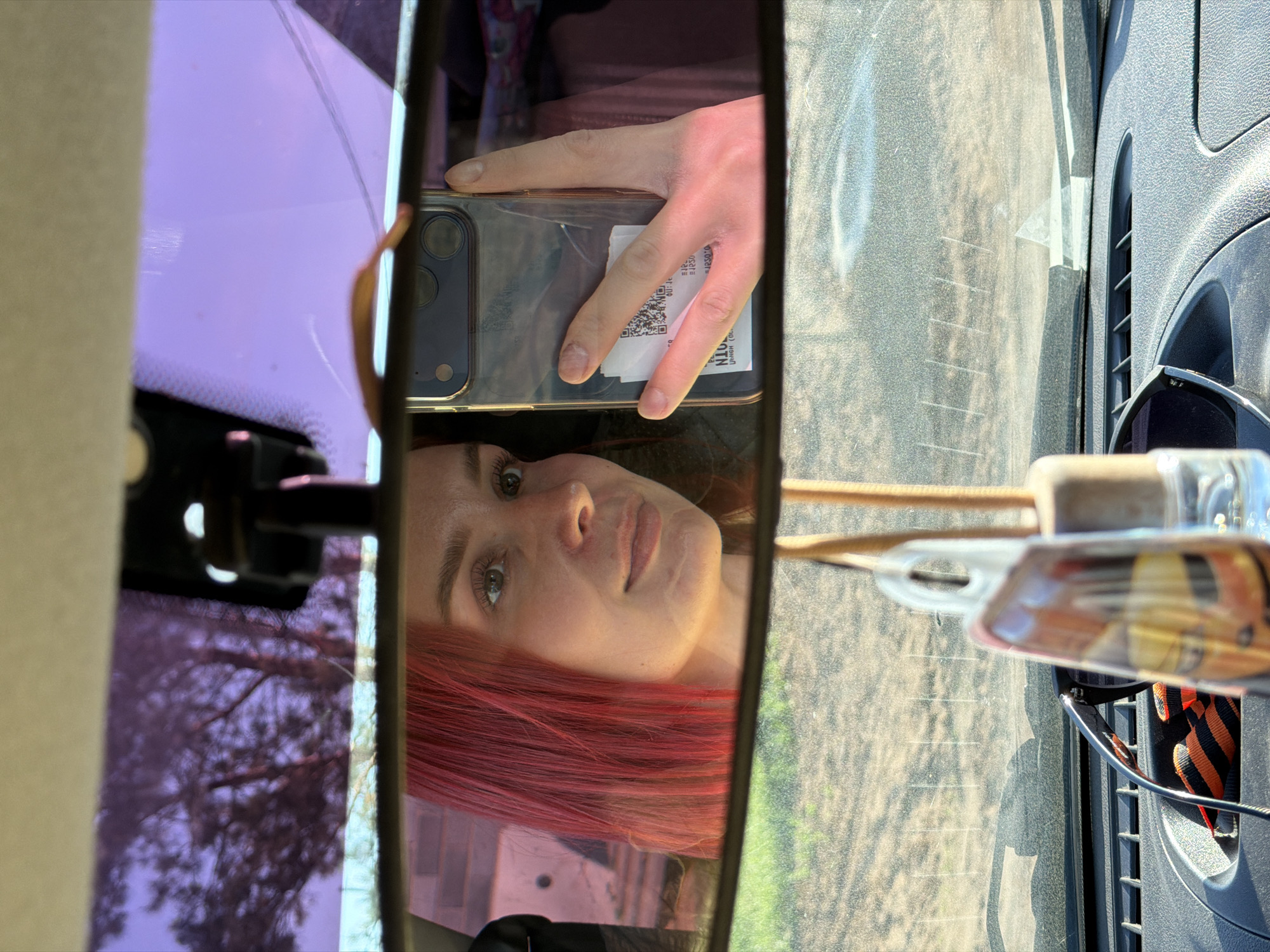 A person with dyed red hair is visible in the rearview mirror of a vehicle. The person is holding a smartphone. The vehicle appears to be in the city of Chaikovsky, Russia, based on text on the smartphone reflecting in the rearview mirror. The image is framed by the rearview mirror and sections of the vehicle interior and windshield.