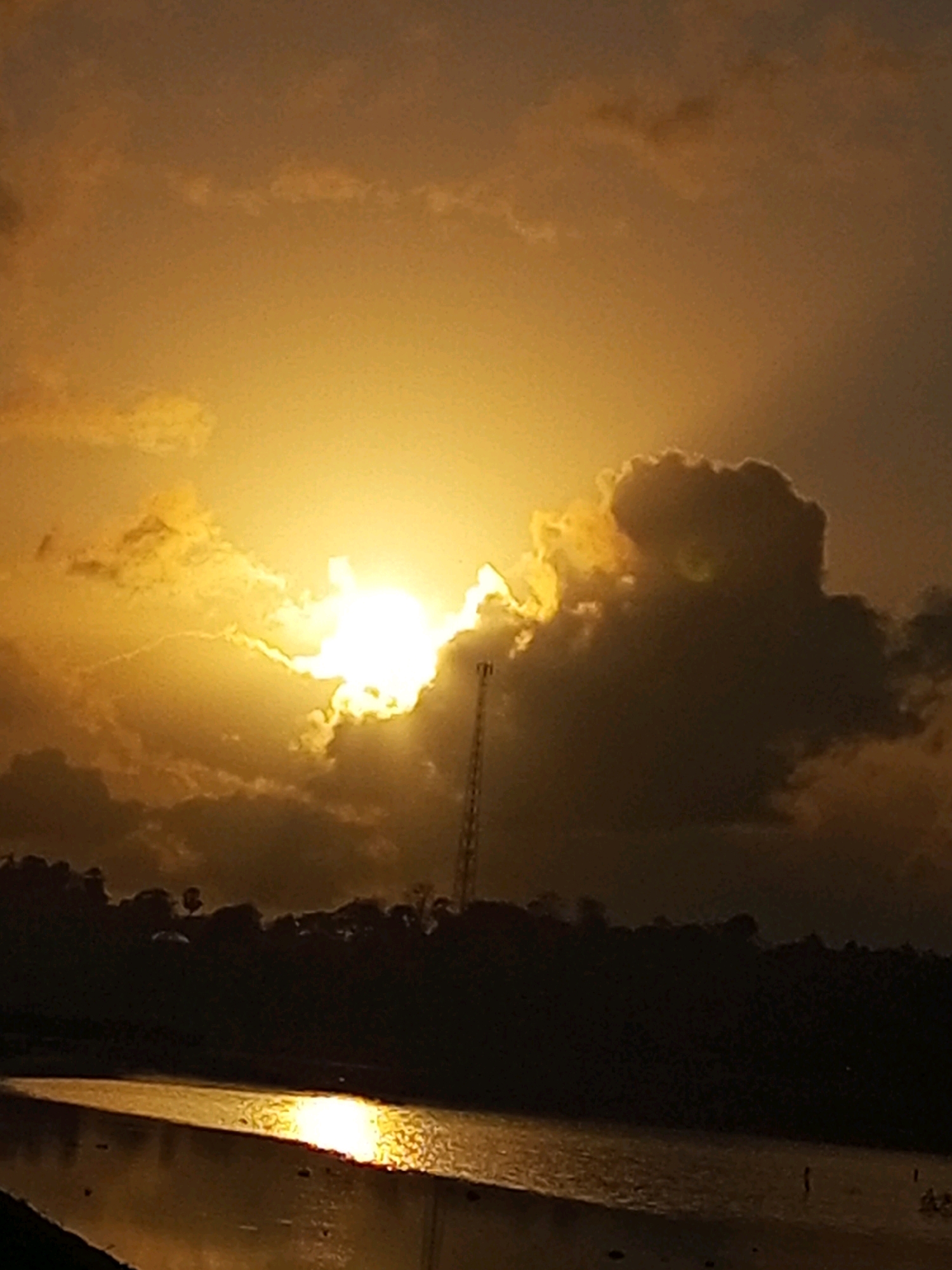 The image depicts a sunset over a body of water, likely a river or lake, near Matasio, Indonesia. The sun is setting on the left side of the frame, casting a golden-orange glow through the clouds and reflecting brightly on the water's surface. A dark silhouette of trees and vegetation lines the far bank of the water. A tall, slender cell tower stands in the middle ground, its dark form contrasting with the vibrant sky. The water itself appears calm, with the reflection of the sunset creating a shimmering effect. There are no discernible individuals or explicit actions taking place within the frame.