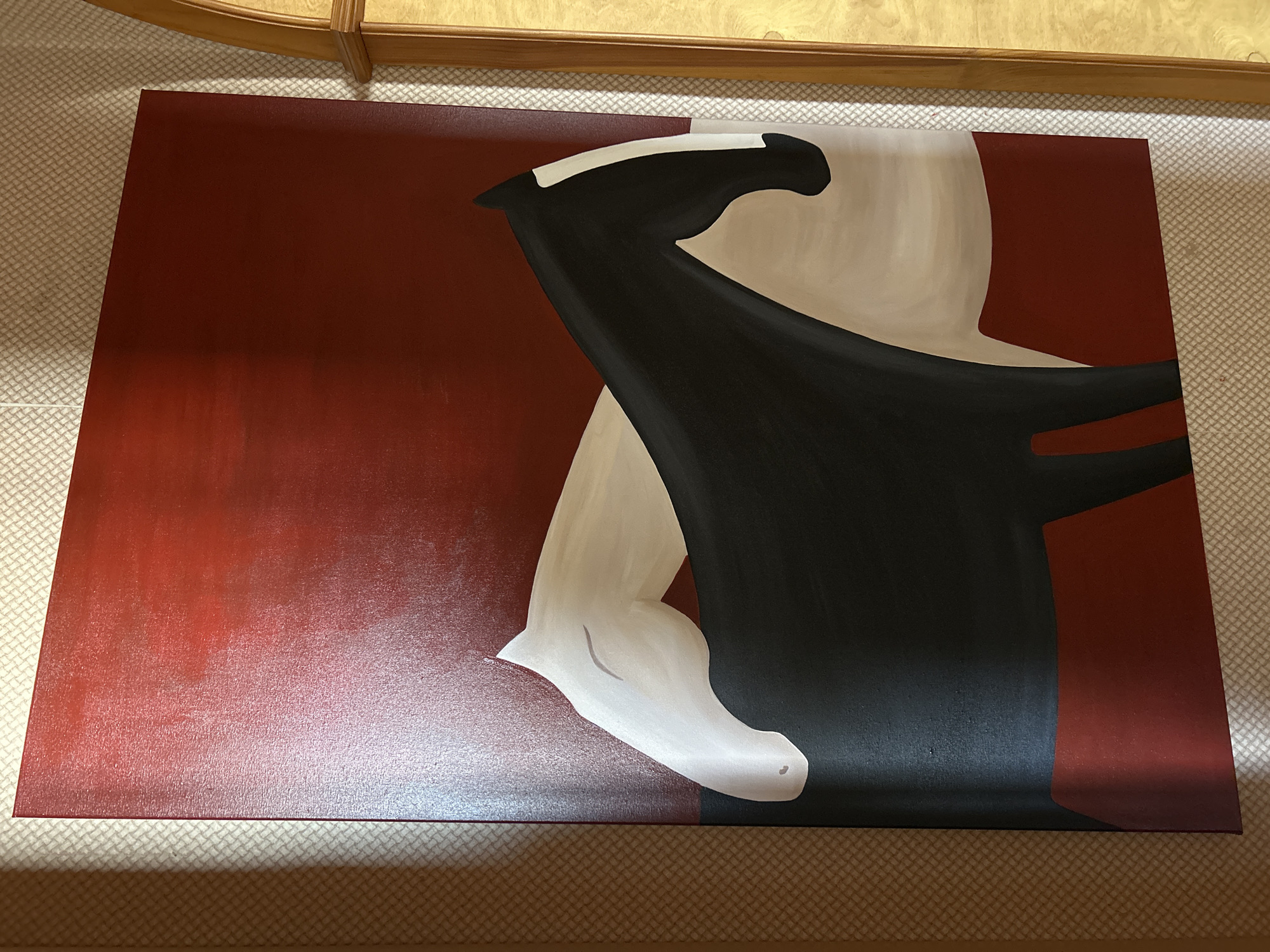 A vertical, close-up, eye-level shot shows a painting of two horses on a textured wall. The painting, predominantly in shades of red, black, white, and beige, depicts two stylized horses facing each other. The horse in the foreground is black with a white stripe on its face and a beige underbelly. The horse behind it is white, with its head and neck curved to nuzzle the black horse. The background of the painting is a gradient of deep red. The painting is hung on a wall with a light, patterned wallpaper. To the right of the painting, a portion of a wooden archway is visible. The scene is lit by an unseen light source from the right, casting shadows on the wall. This image was taken in Moscow, Russia.