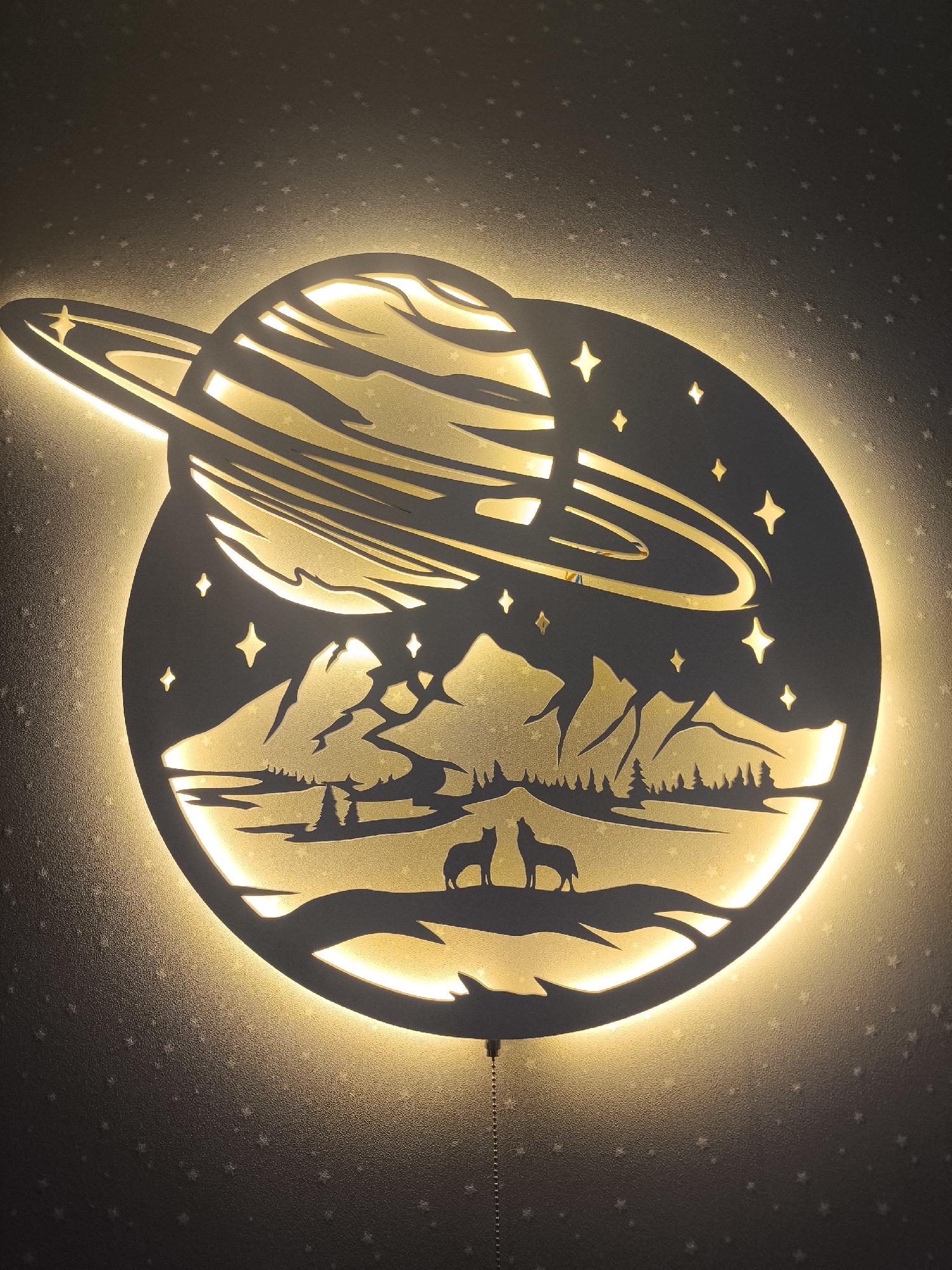 The image shows an illuminated wall fixture depicting a planet with rings, featuring a landscape scene within its silhouette. The landscape includes mountains, trees, and two wolf silhouettes standing on a raised area. The fixture has star cutouts and a pull chain. The wall behind the fixture has a star pattern. The location is not discernible from the image; there is no indication it is Moscow, Russia.